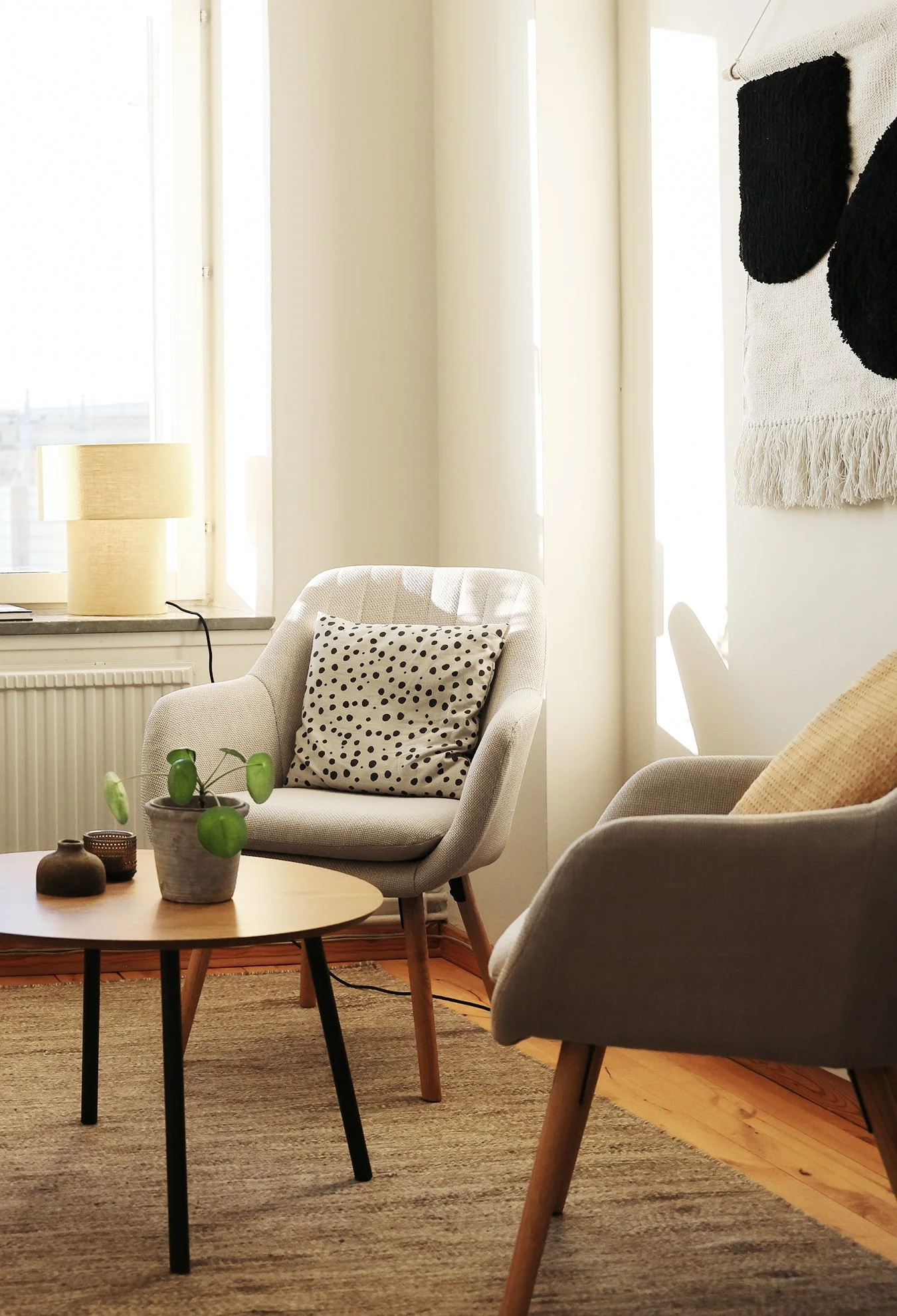 Samtalsrum med två fåtöljer, ett runt soffbord, en växt i kruka, ett fönster med vitt gardin och en väggmålning med svart och beige väv.