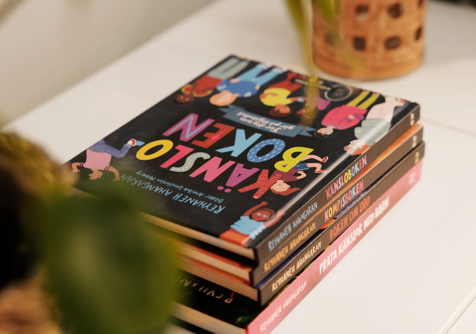 En stapel böcker på ett bord, den översta boken har en svart omslag med färgglada bokstäver och tecknade figurer, titeln är 'Känsloboken'.