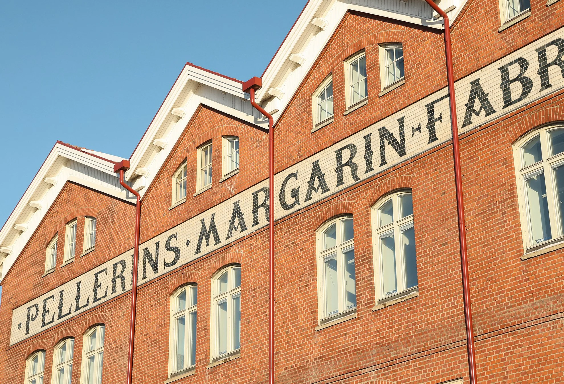 En röd tegelbyggnad med vitt fönsterband och texten 'Pellegrins Margarinfabrik' på sidan, under ett klart blå himmel.