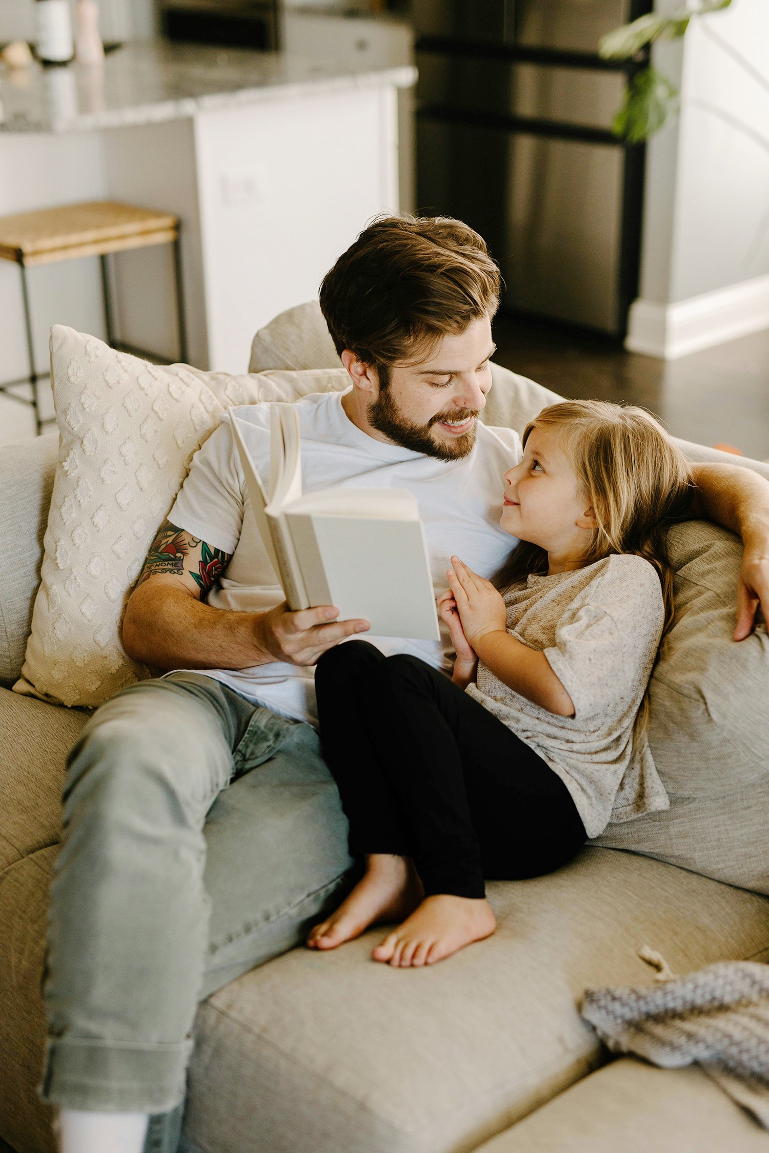 En förälder och ett barn som tittar på varandra och läser en bok tillsammans på en soffa i ett hem.