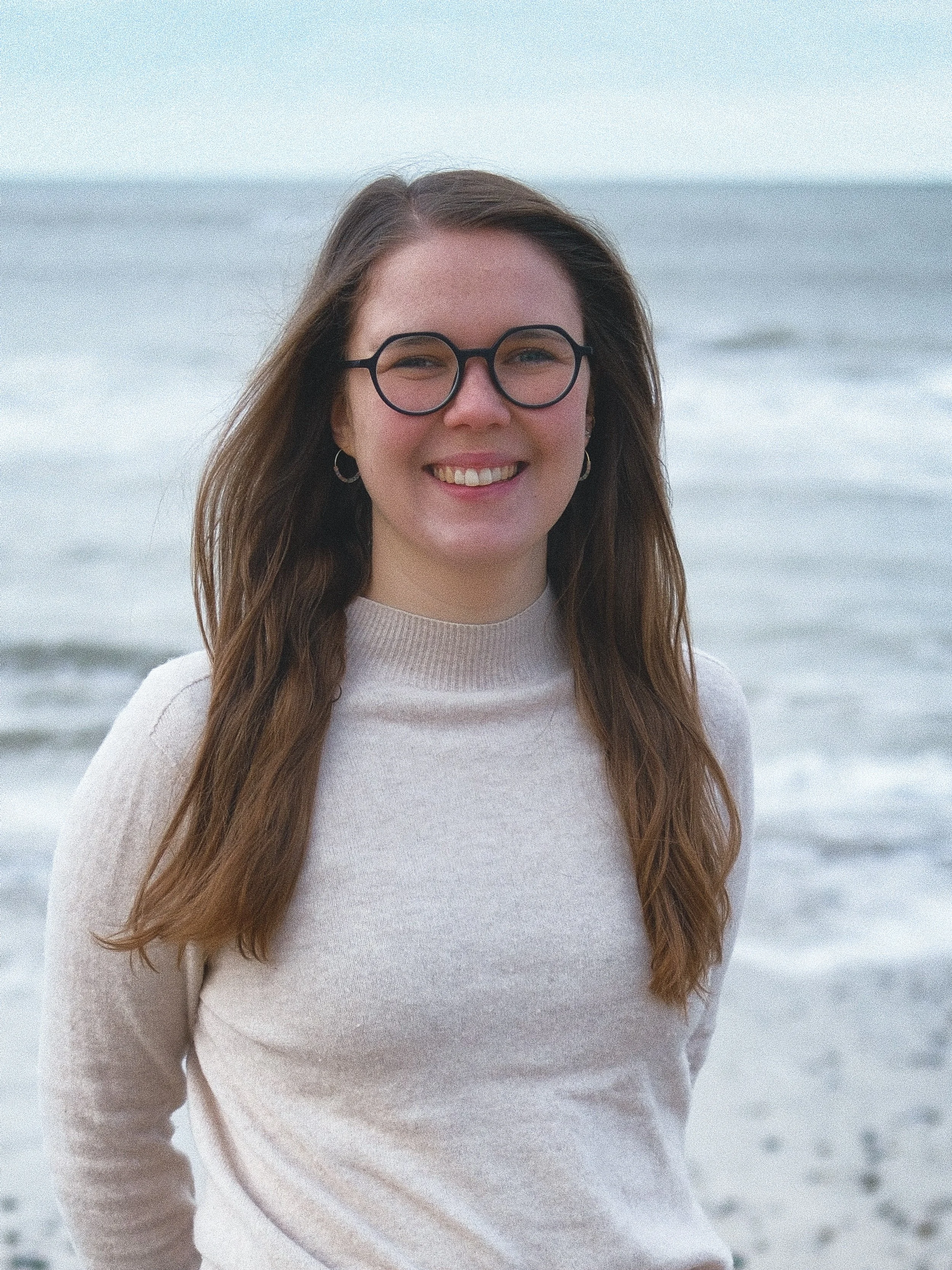 Junge Frau mit Brille und lockigem braunem Haar beim Strand, lächelnd in die Kamera schauend.