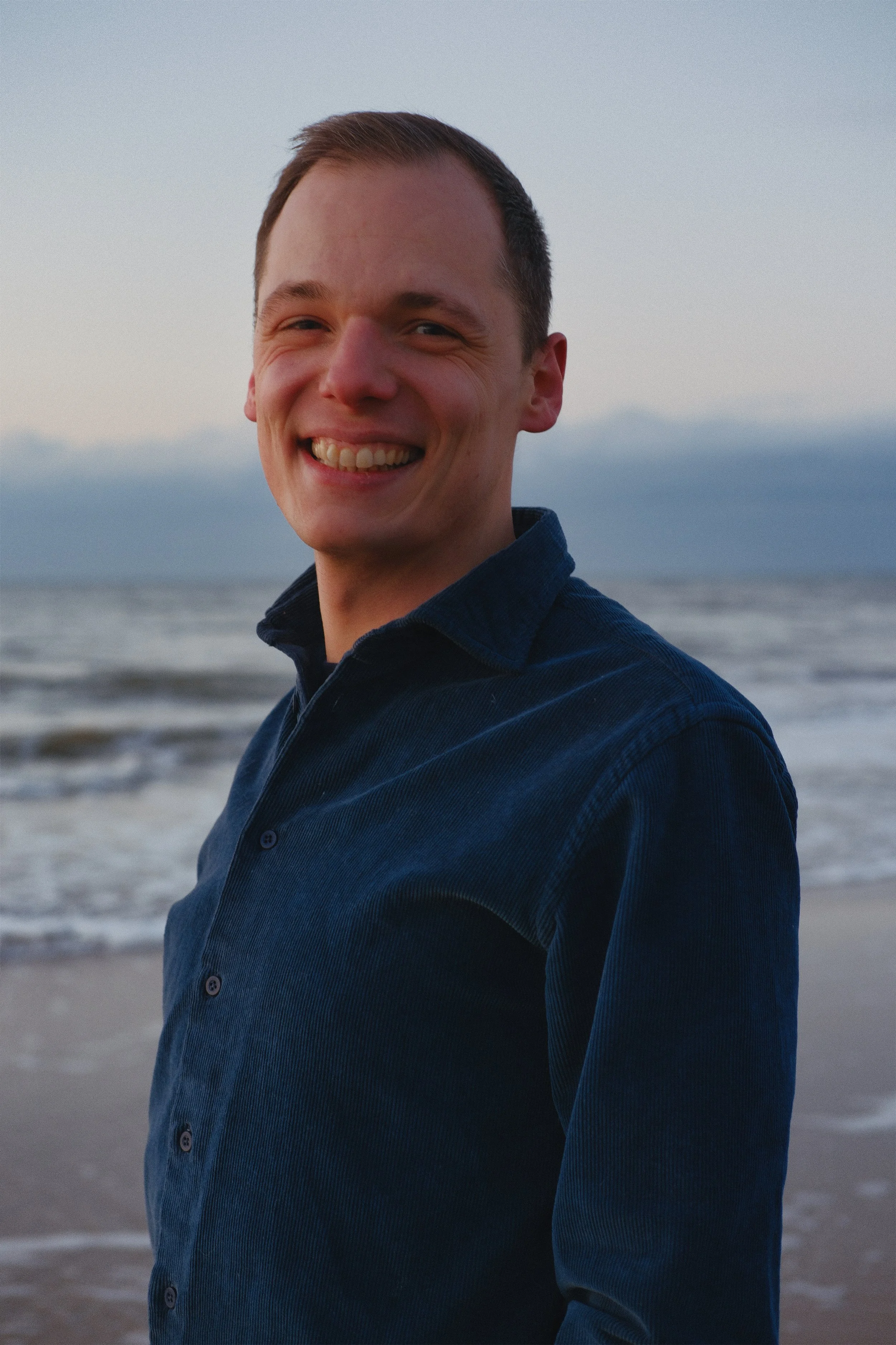 Ein lachender Mann im Dunkelblauen Hemd steht am Strand bei sunset, im Hintergrund das Meer und Himmel.