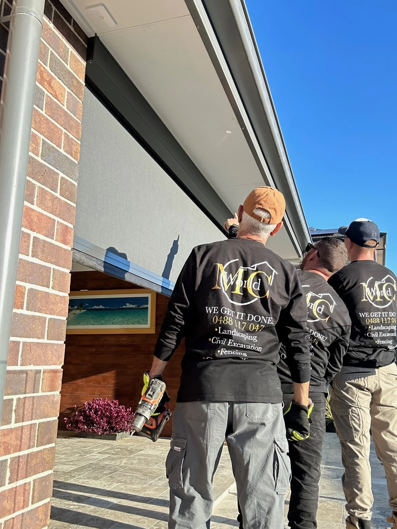 Three workers in black shirts with company logo inspecting a window outside a building with brick wall and flowering plants.