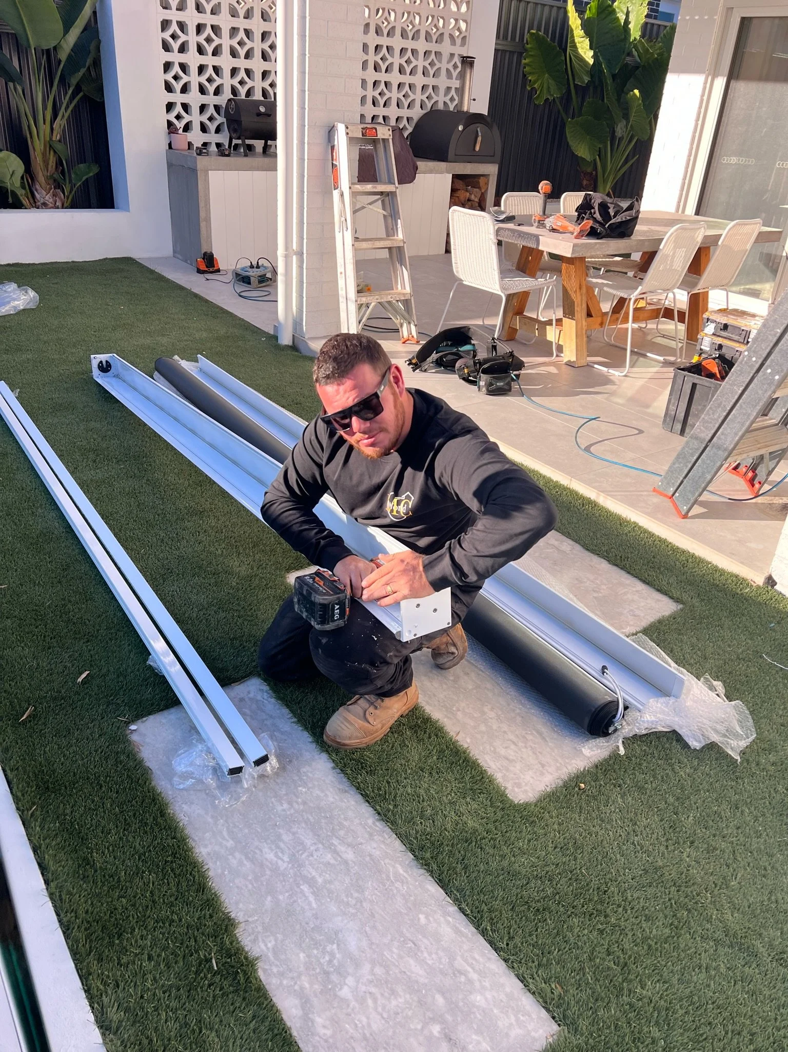 A man kneels on artificial grass holding a power drill and working with metal beams outdoors, with construction tools and materials scattered around.