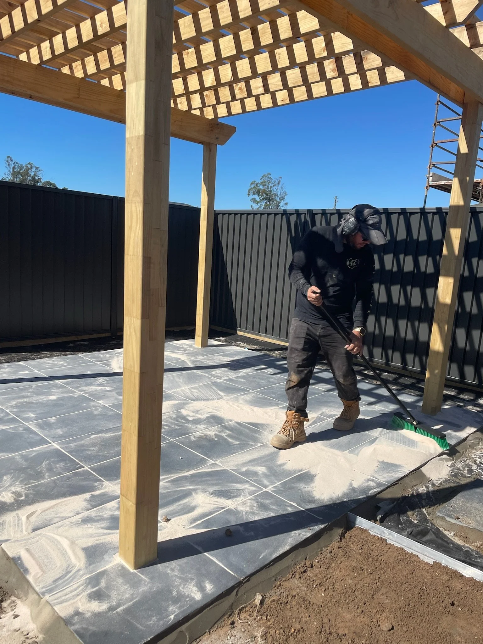 A construction worker raking white powdery substance on a tiled patio outside a wooden structure under construction, with a black fence and clear blue sky in the background.