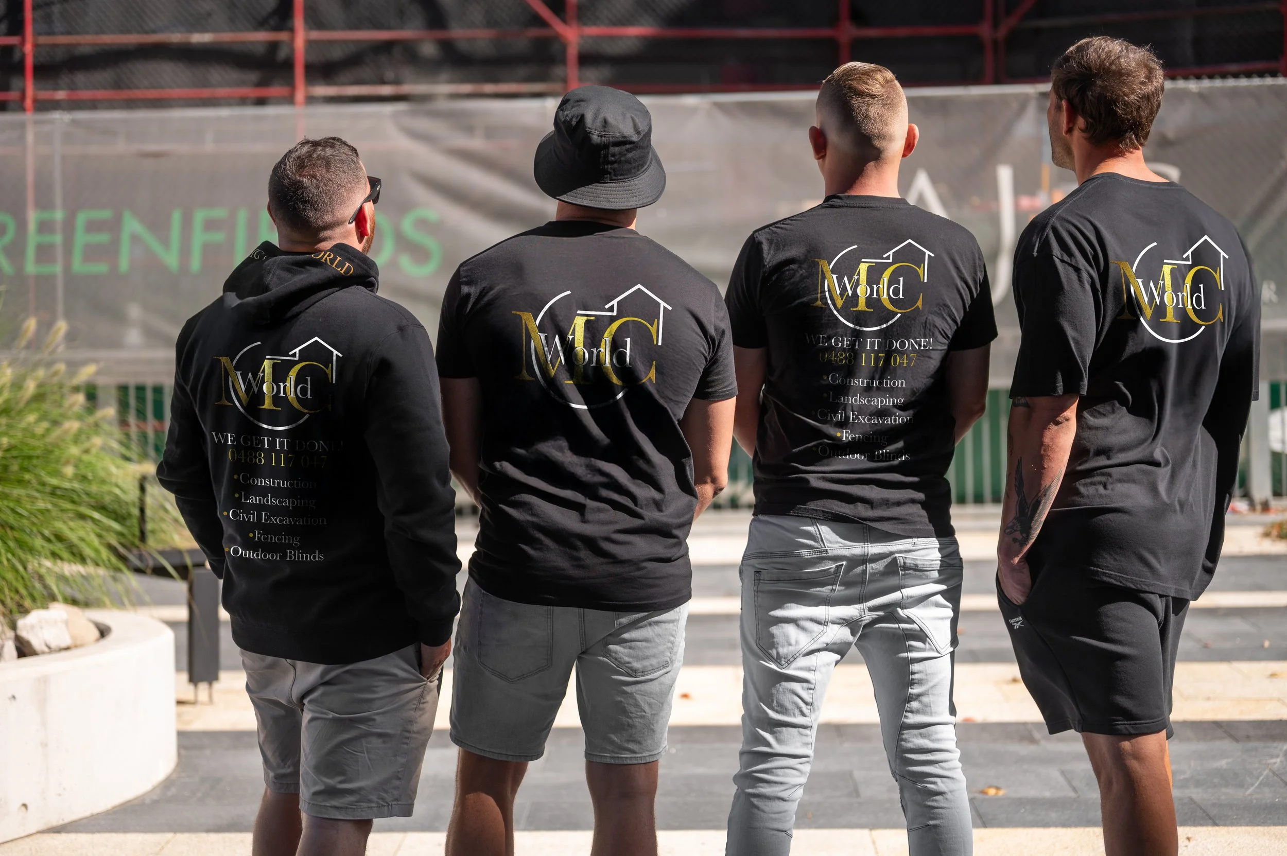 Four men standing outside a sydney based construction site, wearing black T-shirts with a logo and text on the back, facing away from the camera.