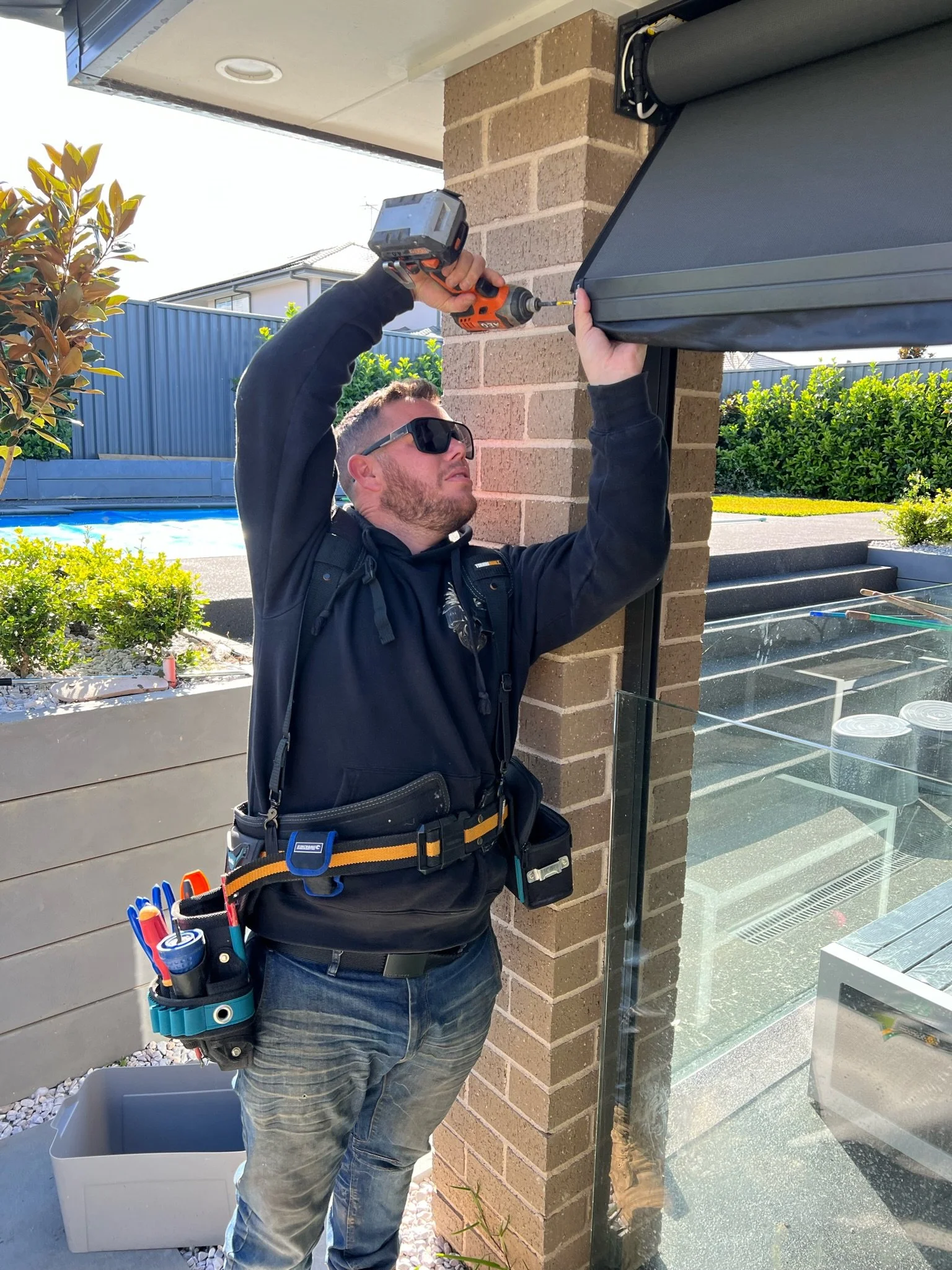 A man wearing sunglasses, a black hoodie, and a tool belt is installing or repairing a window awning with a cordless drill on a brick exterior wall.