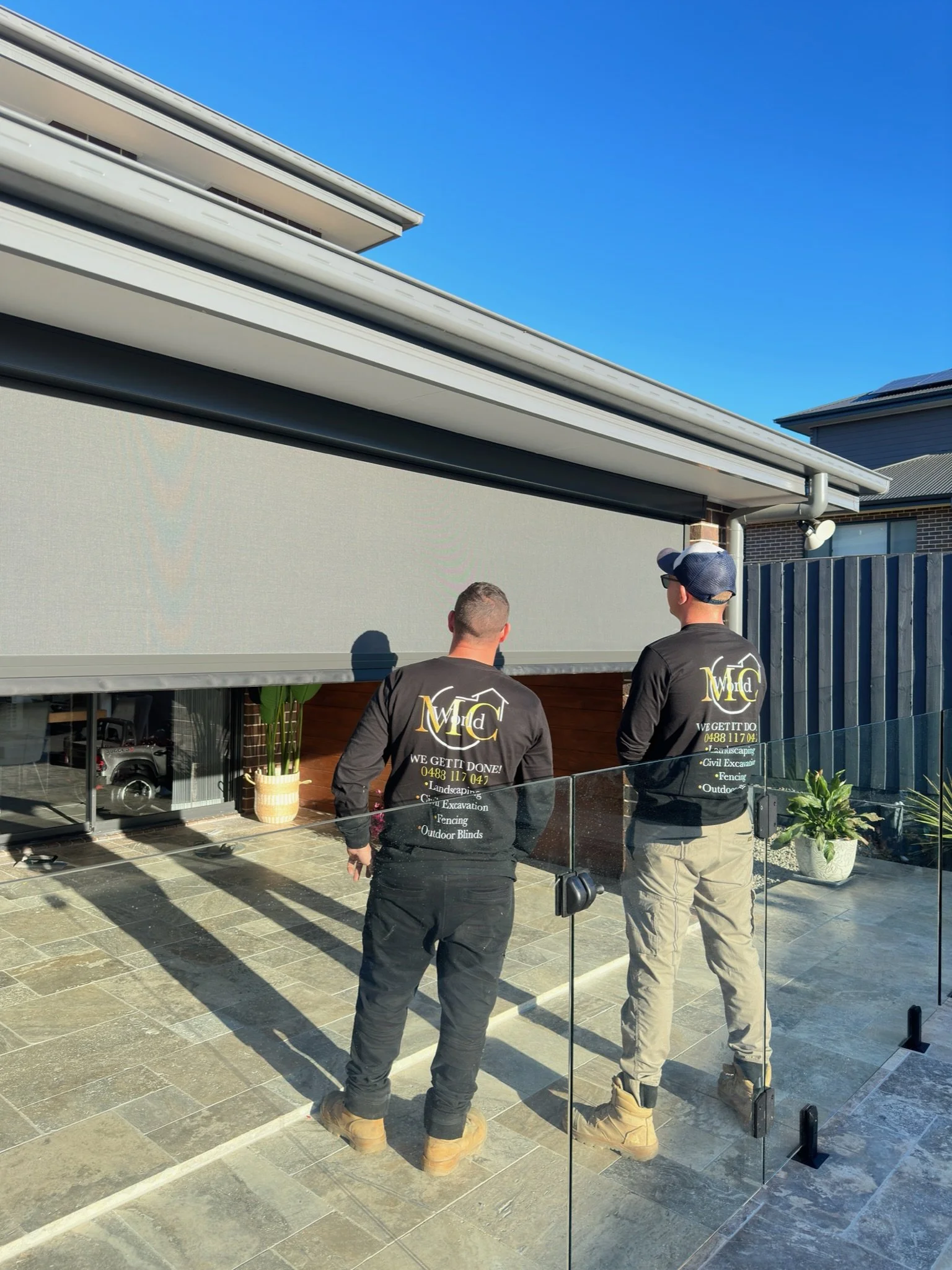 Two workers in black shirts and tan work boots standing outside near a glass fence, facing a large outdoor retractable screen or shade, with a modern house and clear blue sky in the background.