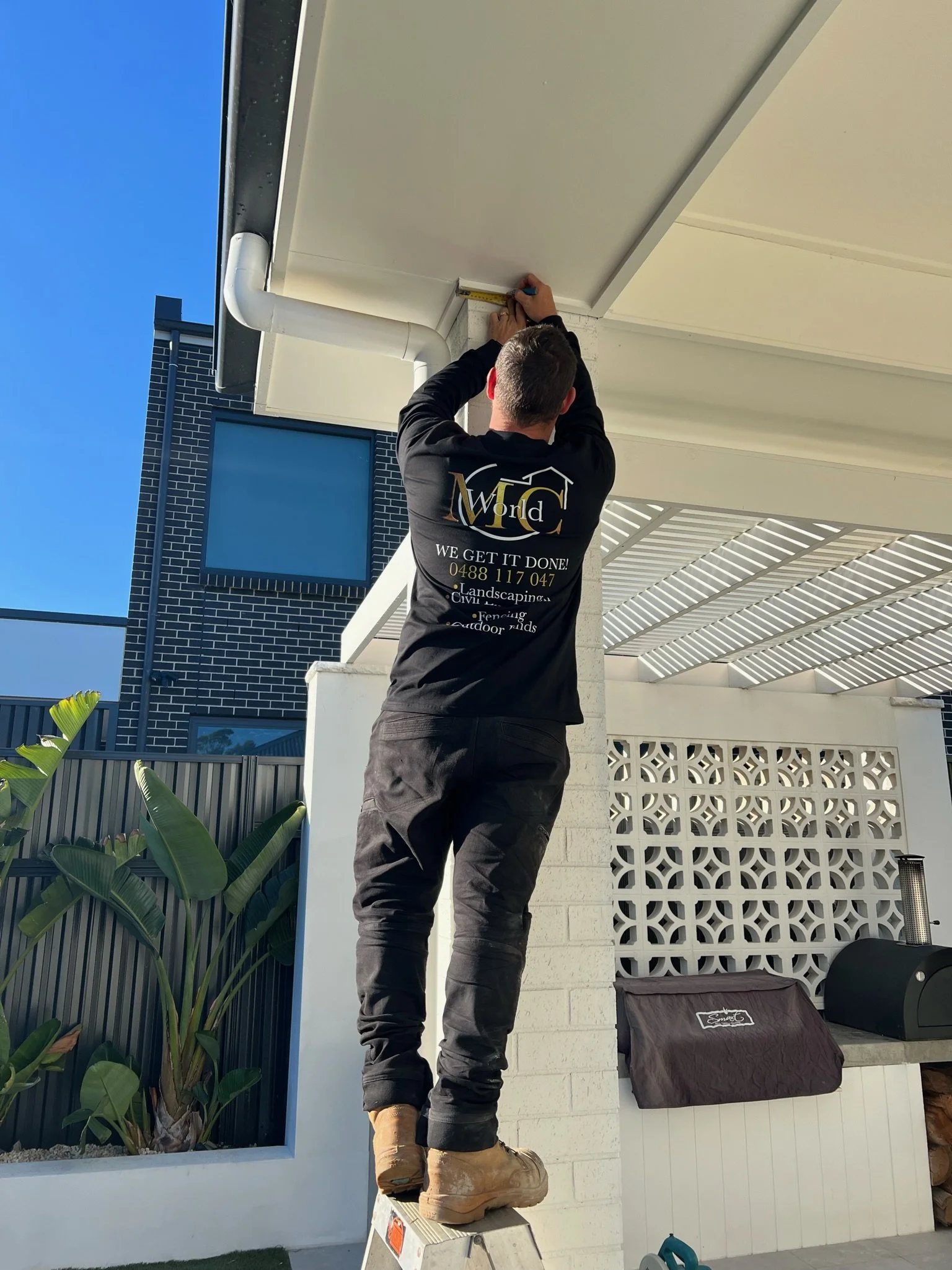 A worker standing on a small ladder measuring the corner of a building's ceiling with a measuring tape.