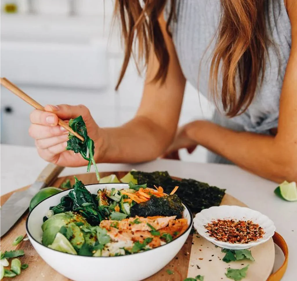 Person using chopsticks to pick up leafy greens from a bowl of salad on a cutting board in a kitchen.