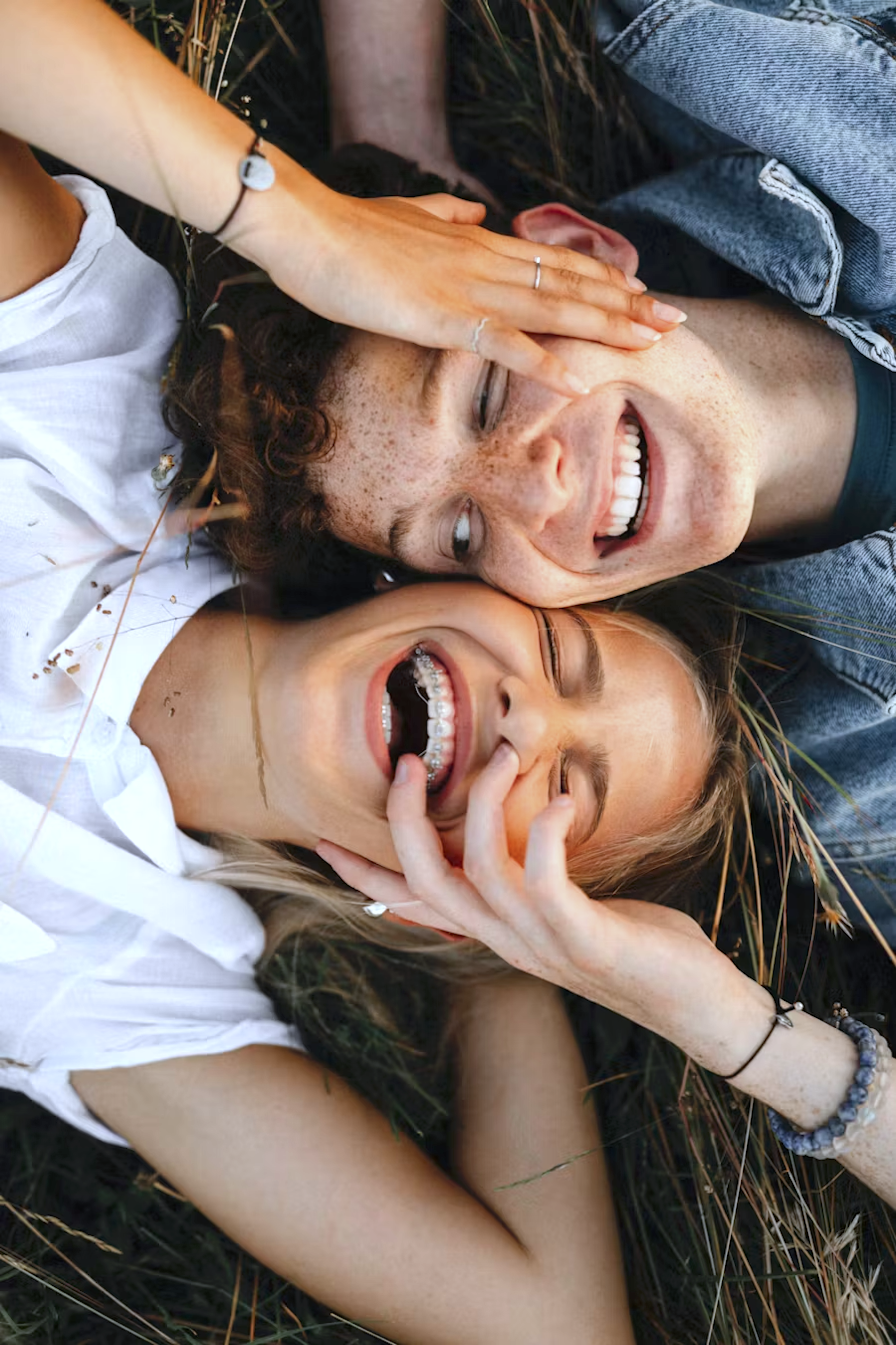 Two smiling women lying on the ground with tall grass, one with red hair and freckles, the other with blonde hair, sharing a joyful moment.