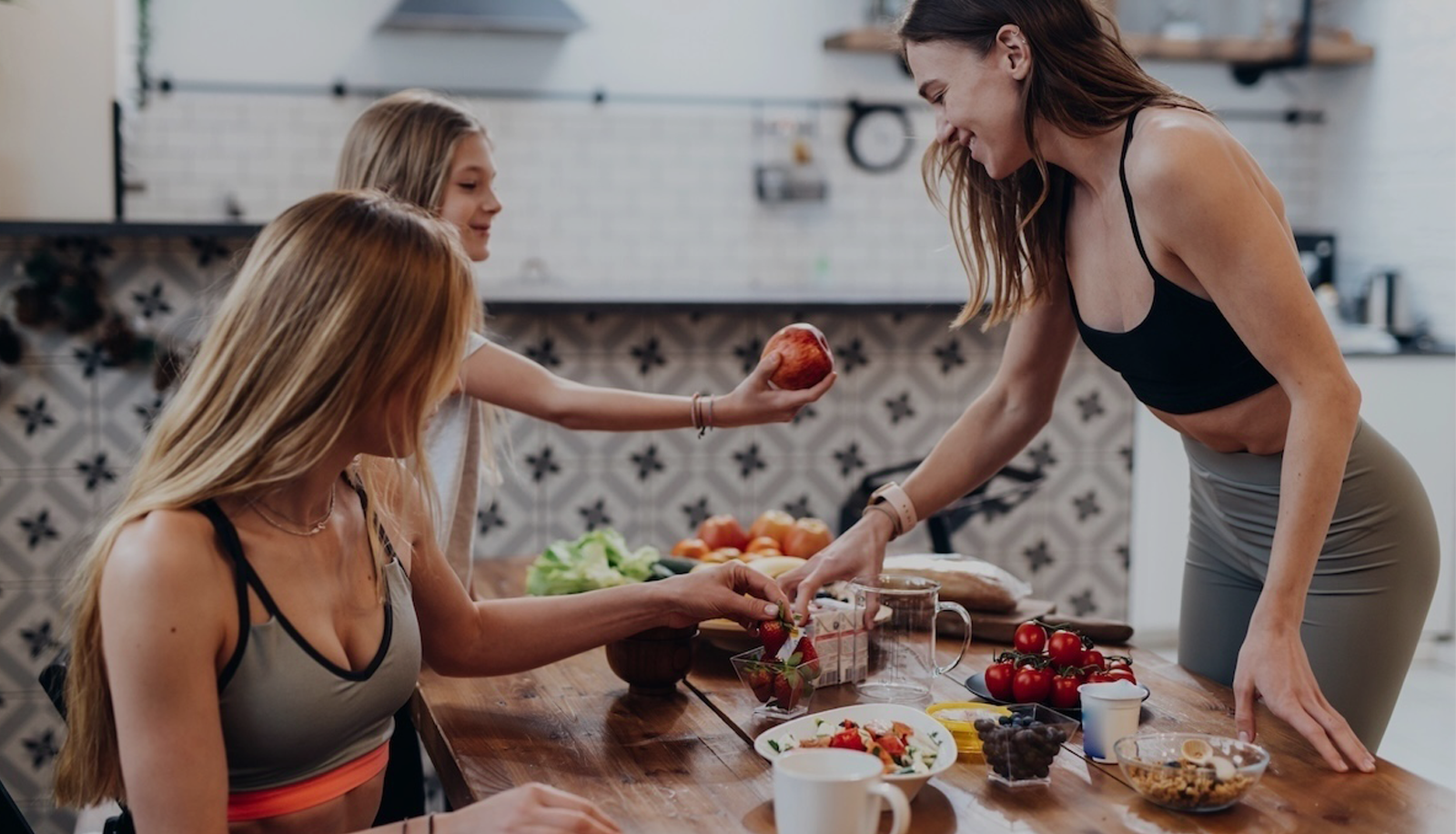 Four women preparing food together in a kitchen, with one woman handing an apple to another, and various fresh fruits on the table.
