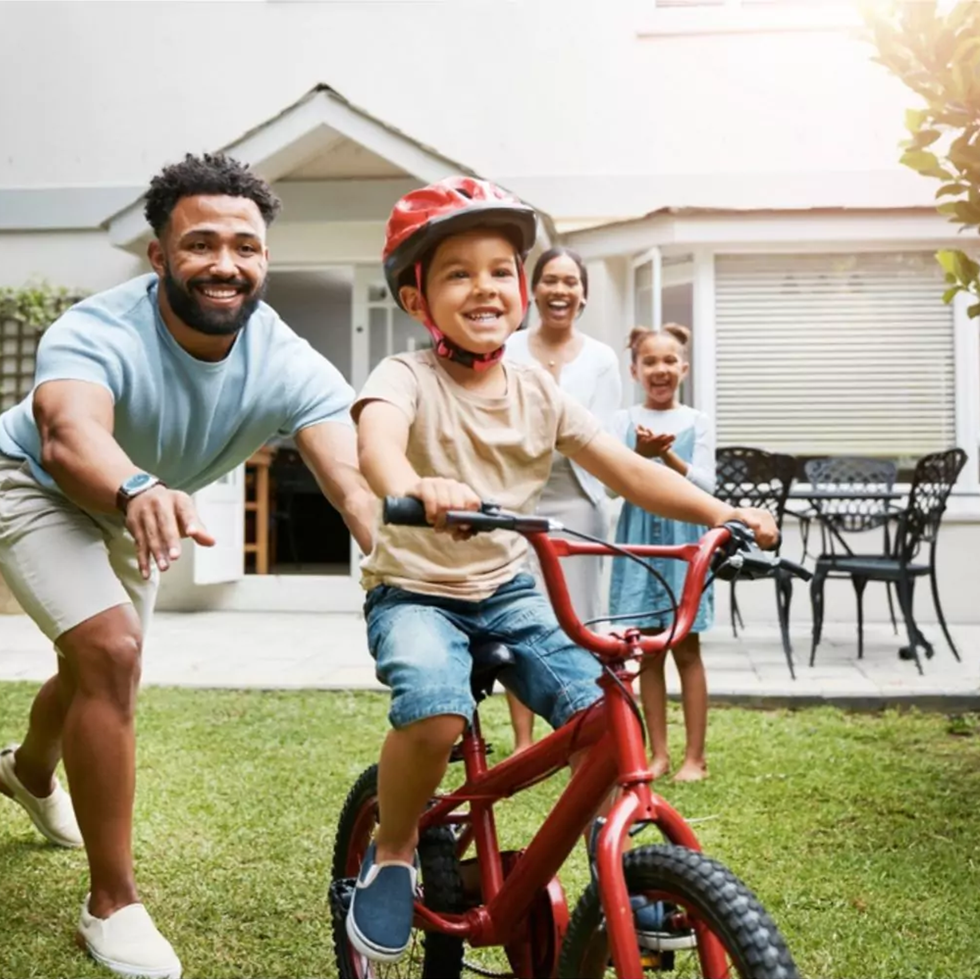 A happy family playing outside in their backyard, with a young boy riding a red bicycle while his family cheers him on.