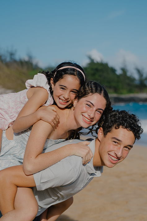 A happy family of three at the beach, with a girl sitting on her dad's back and both smiling, with ocean and sky in the background.