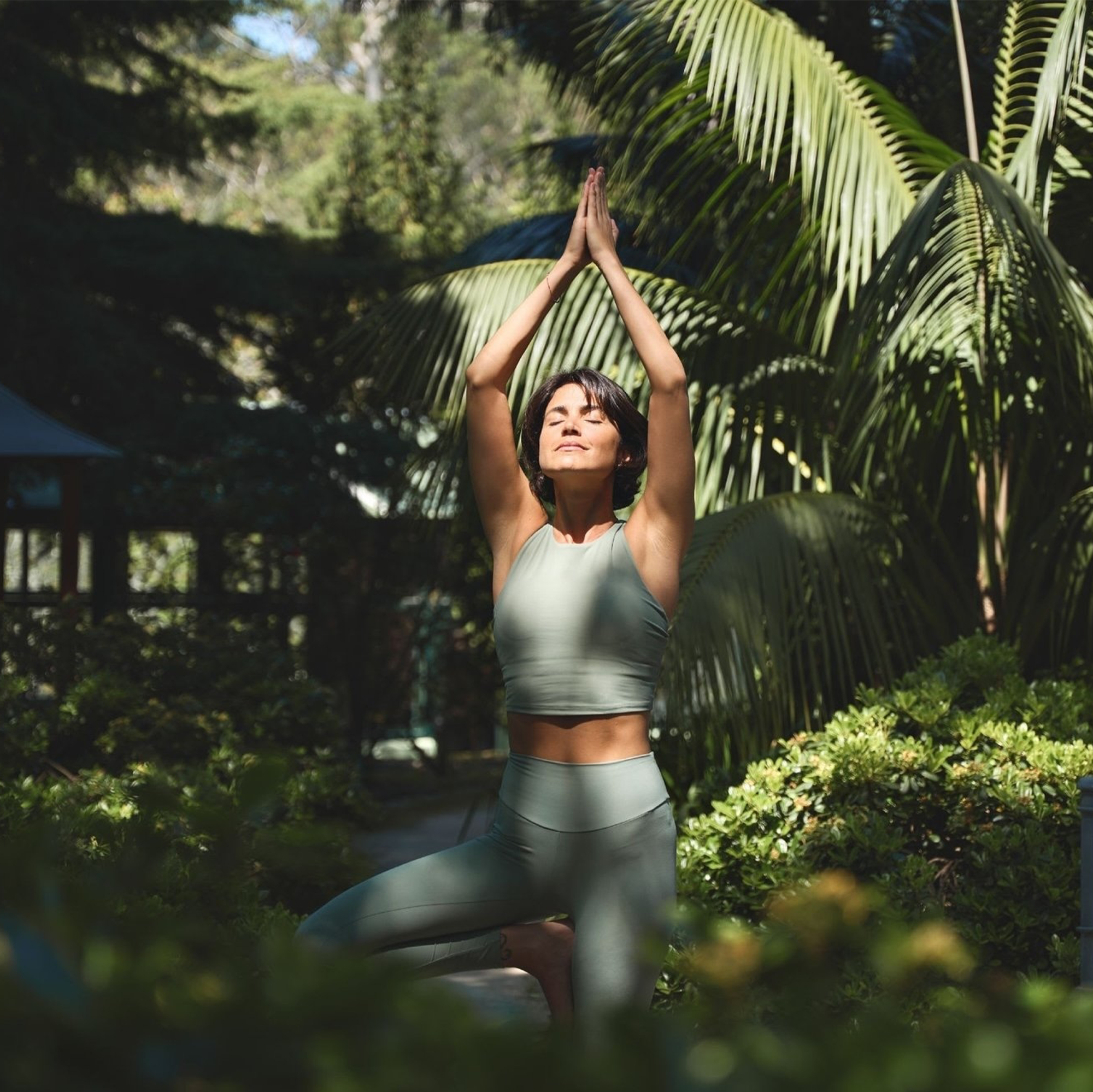 A woman practicing yoga outdoors, standing in a tree pose with hands pressed together above her head, surrounded by lush green plants and palm leaves, with sunlight filtering through the trees.