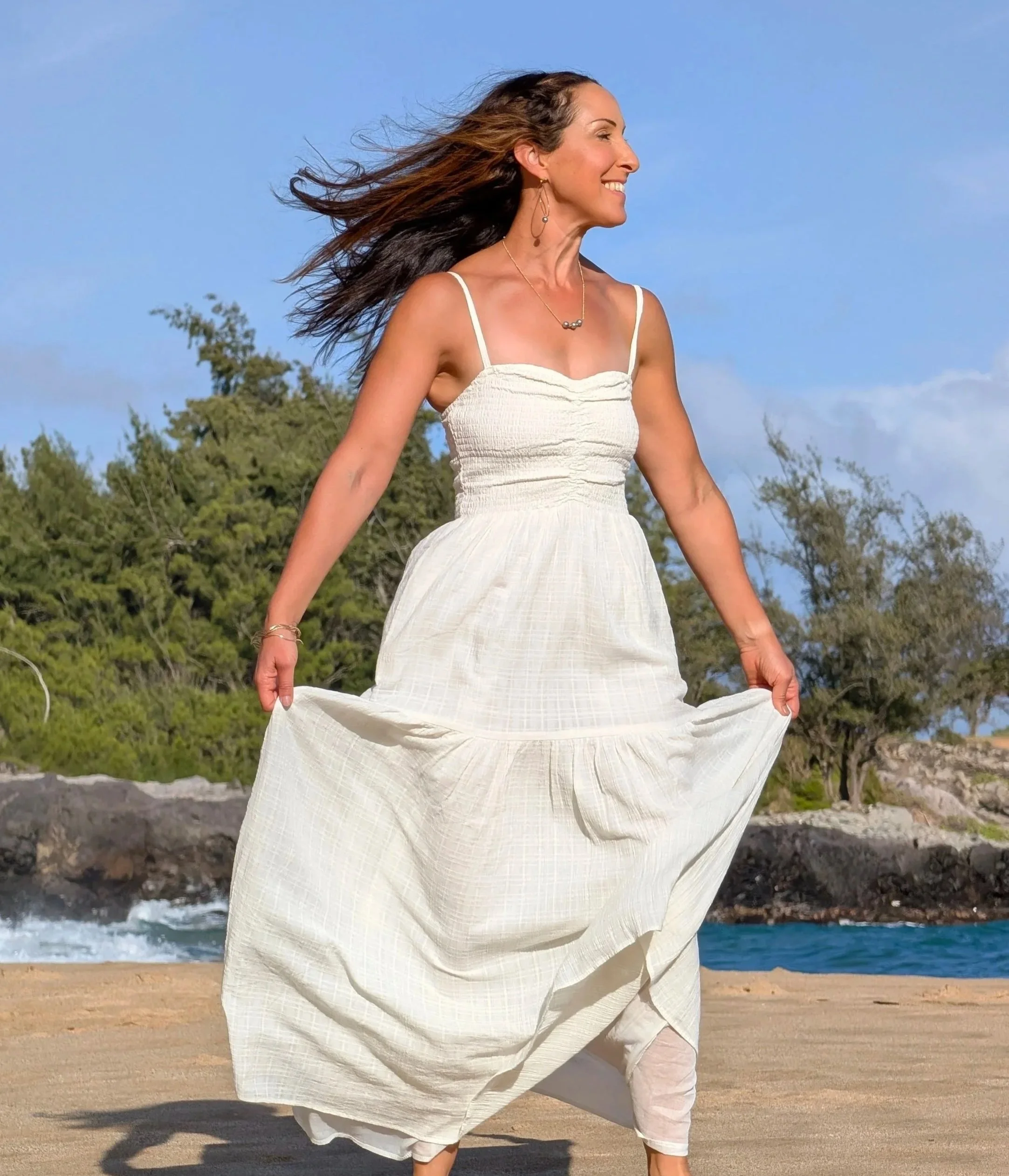 A woman in a white dress smiling and holding the hem of her dress, standing on a sandy beach with rocks, trees, and ocean in the background under a partly cloudy sky.