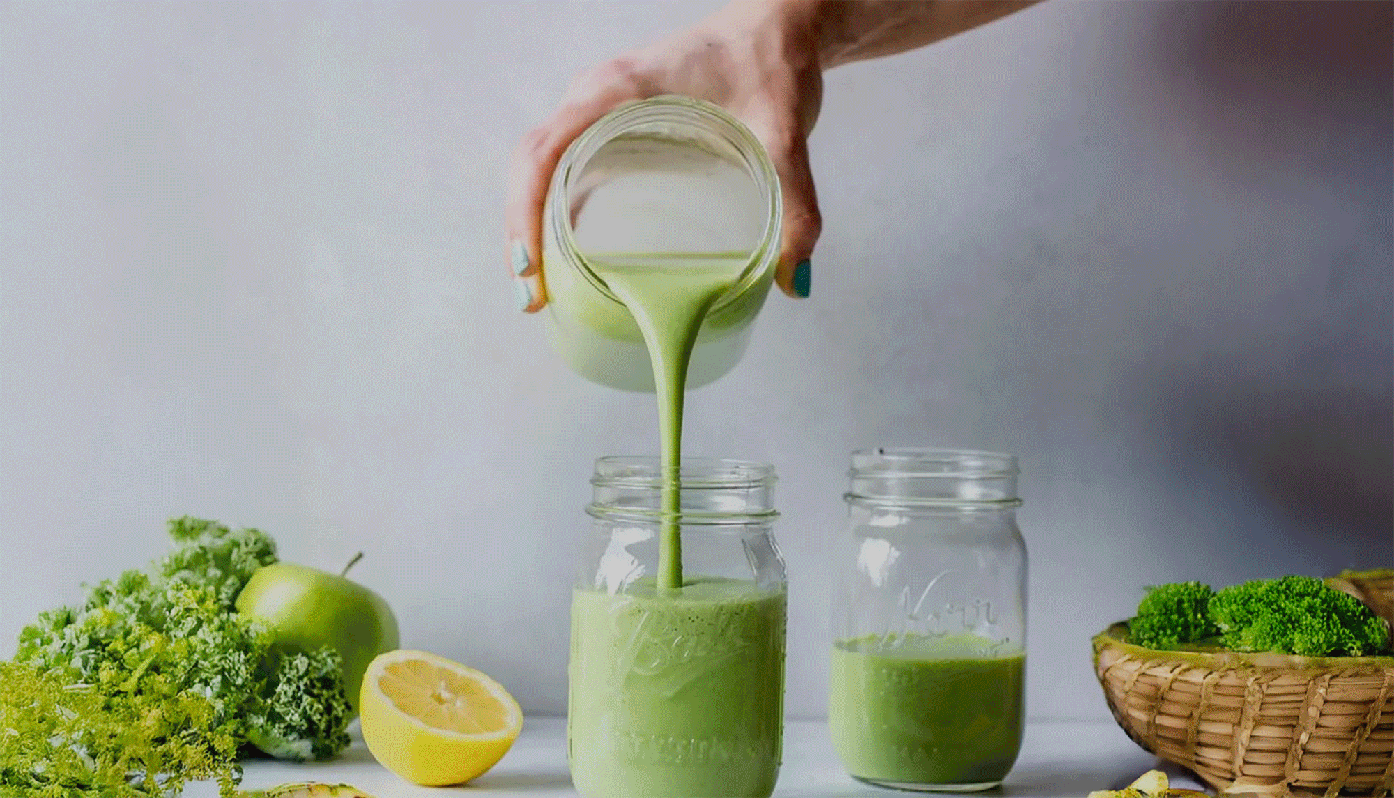 A person pouring green smoothie from a jar into a glass jar, with fresh broccoli, lemon, apple, and other greens on the table.