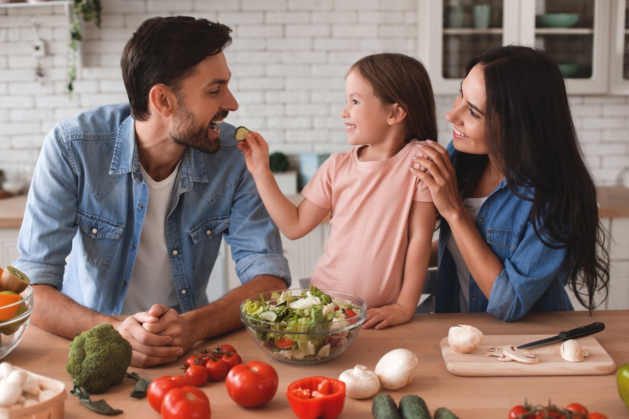 A family of three with a father, mother, and daughter preparing a salad in a kitchen. The father is smiling as the daughter feeds him a slice of cucumber. The mother is smiling and holding the daughter. The counter has tomatoes, broccoli, mushrooms, and a bowl of salad.