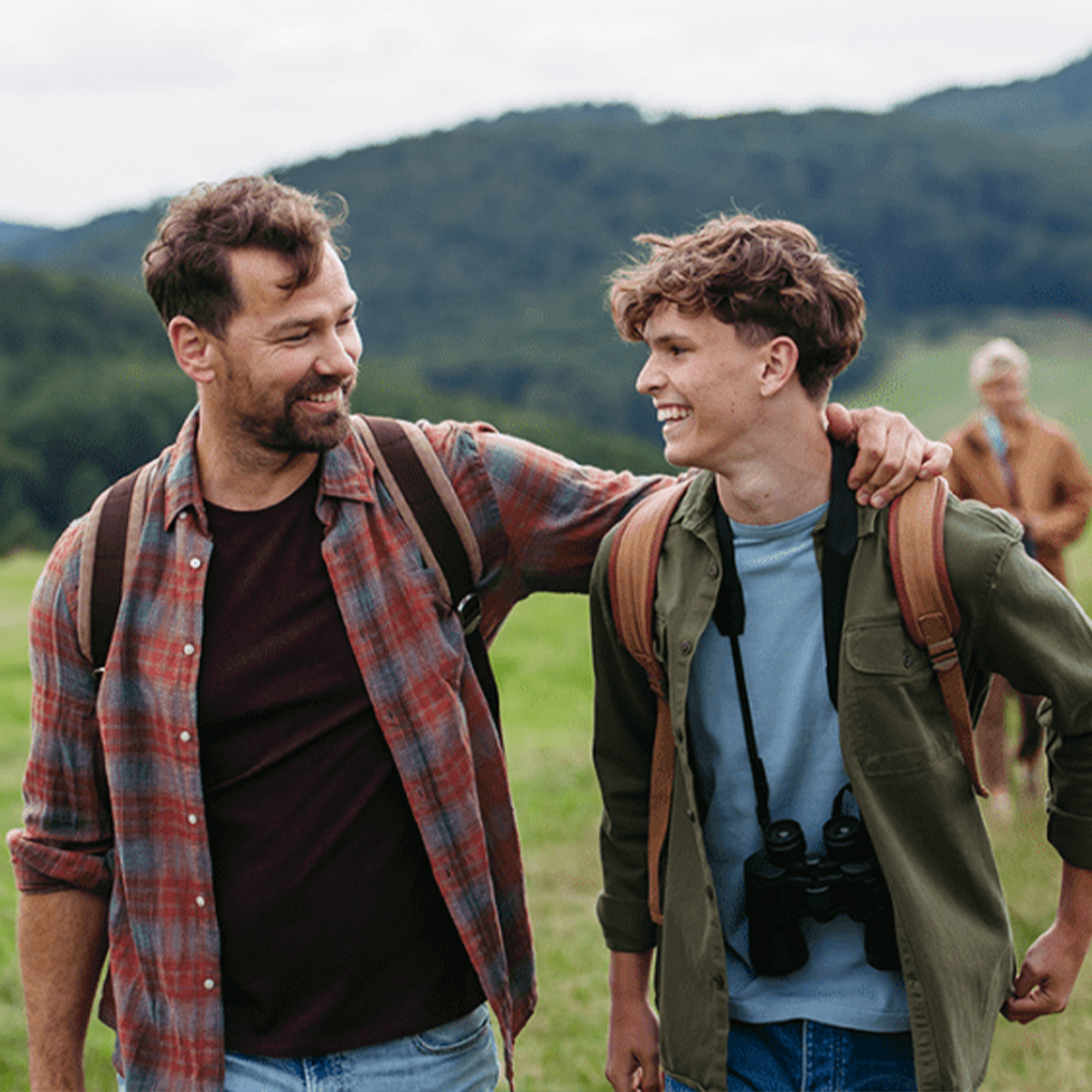 Two men, one with a beard and wearing a plaid shirt, and the other young with curly hair and a camera around his neck, smiling and walking outdoors in a green, mountainous landscape with another person in the background.