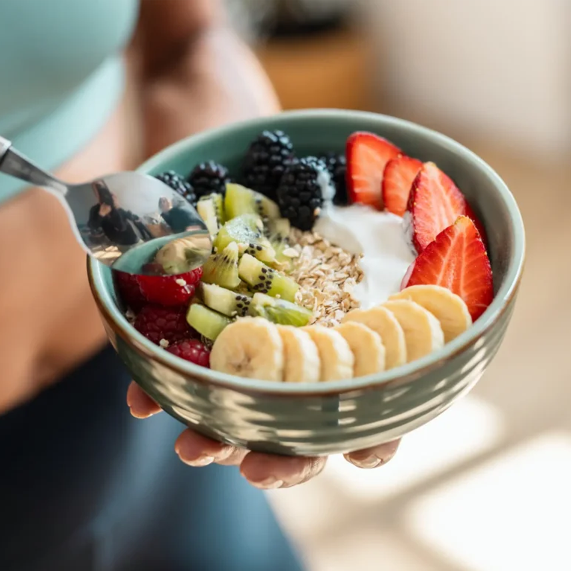 A person holding a bowl of yogurt topped with strawberries, bananas, kiwi, blackberries, raspberries, granola, and oats.