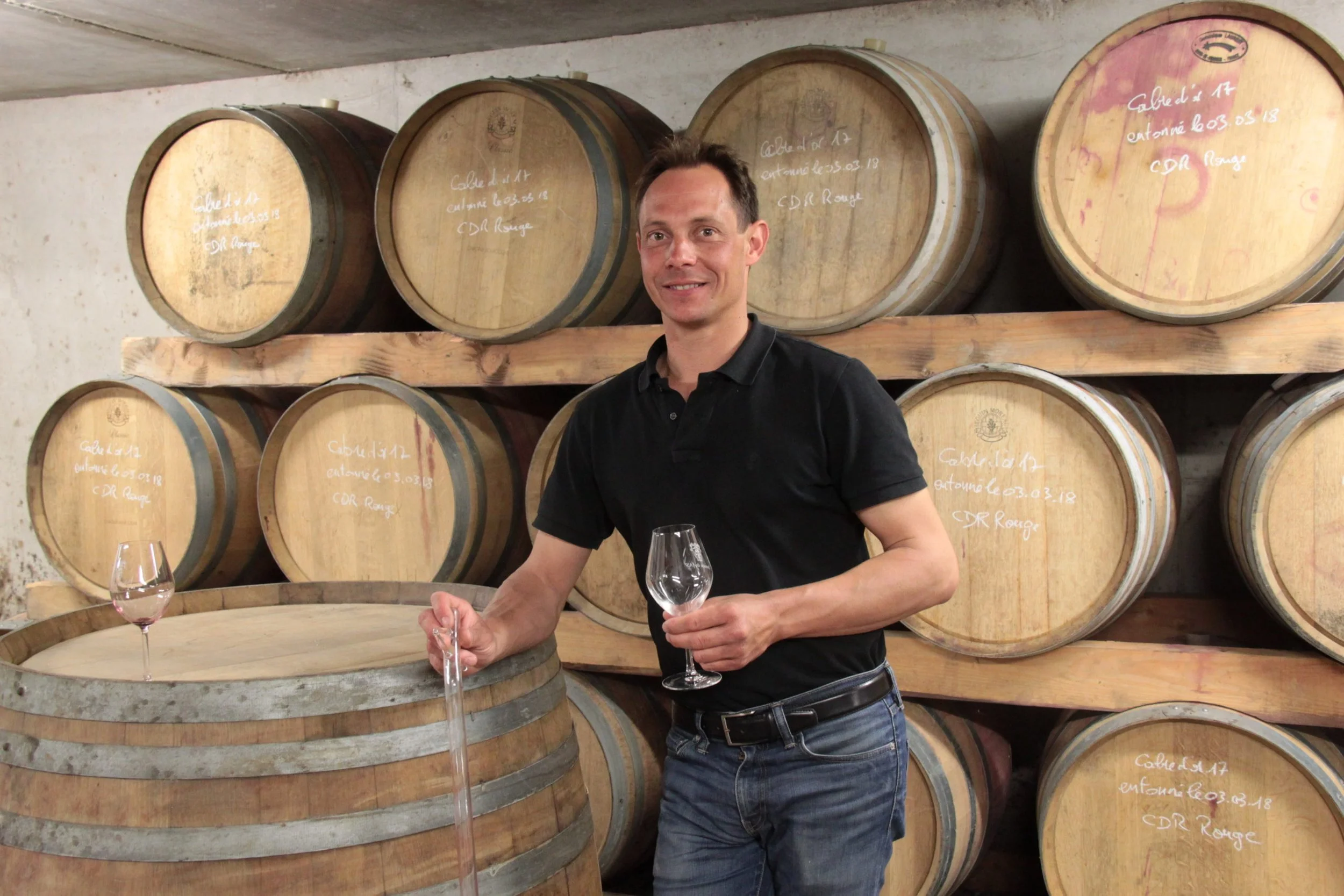 Un homme en polo noir et jeans, tenant un verre de vin, debout devant des barriques en bois dans une cave à vin.