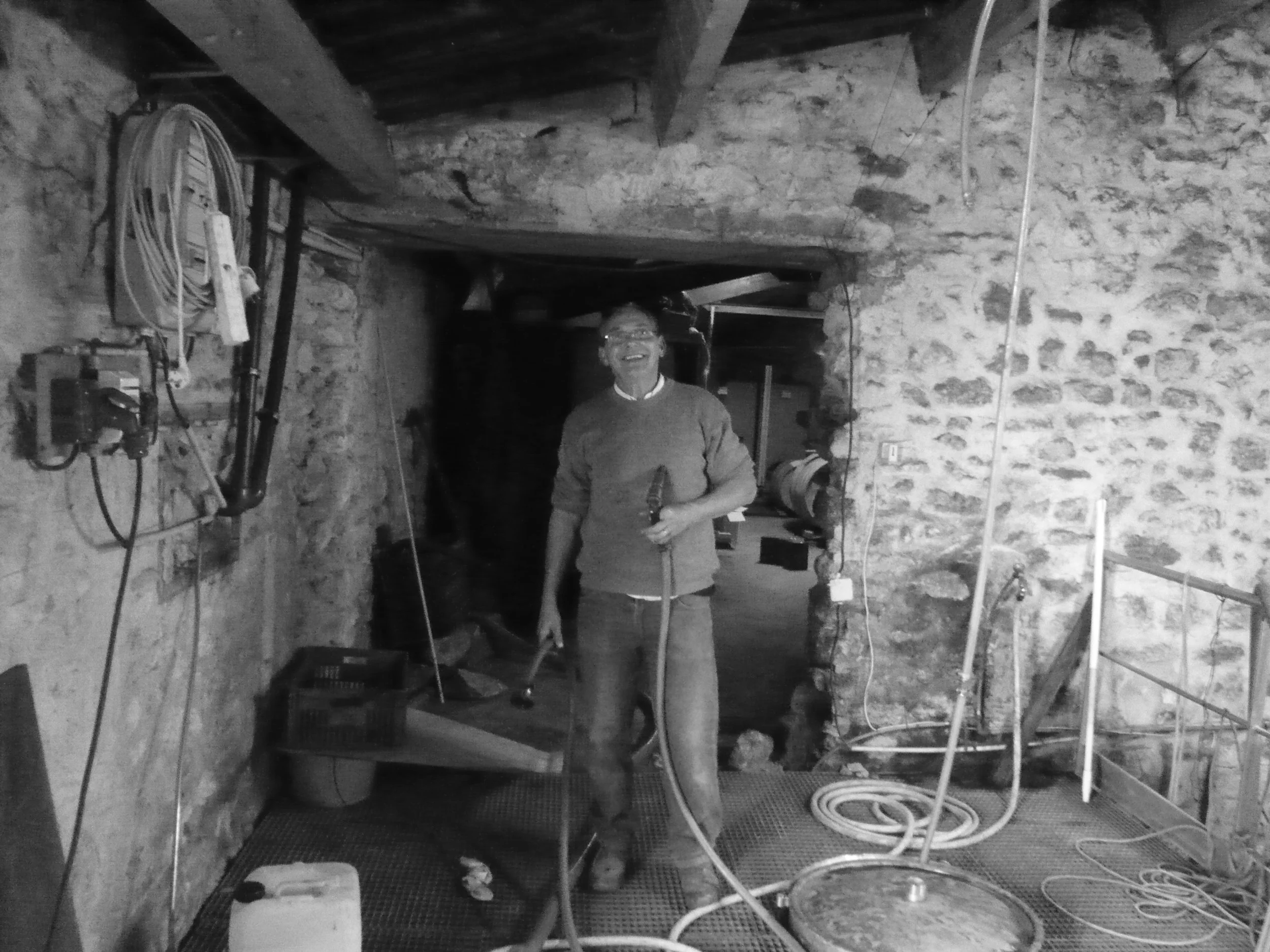 Un homme souriant tenant un outil dans un espace de travail avec des murs en pierre, équipement électrique et câbles suspendus.