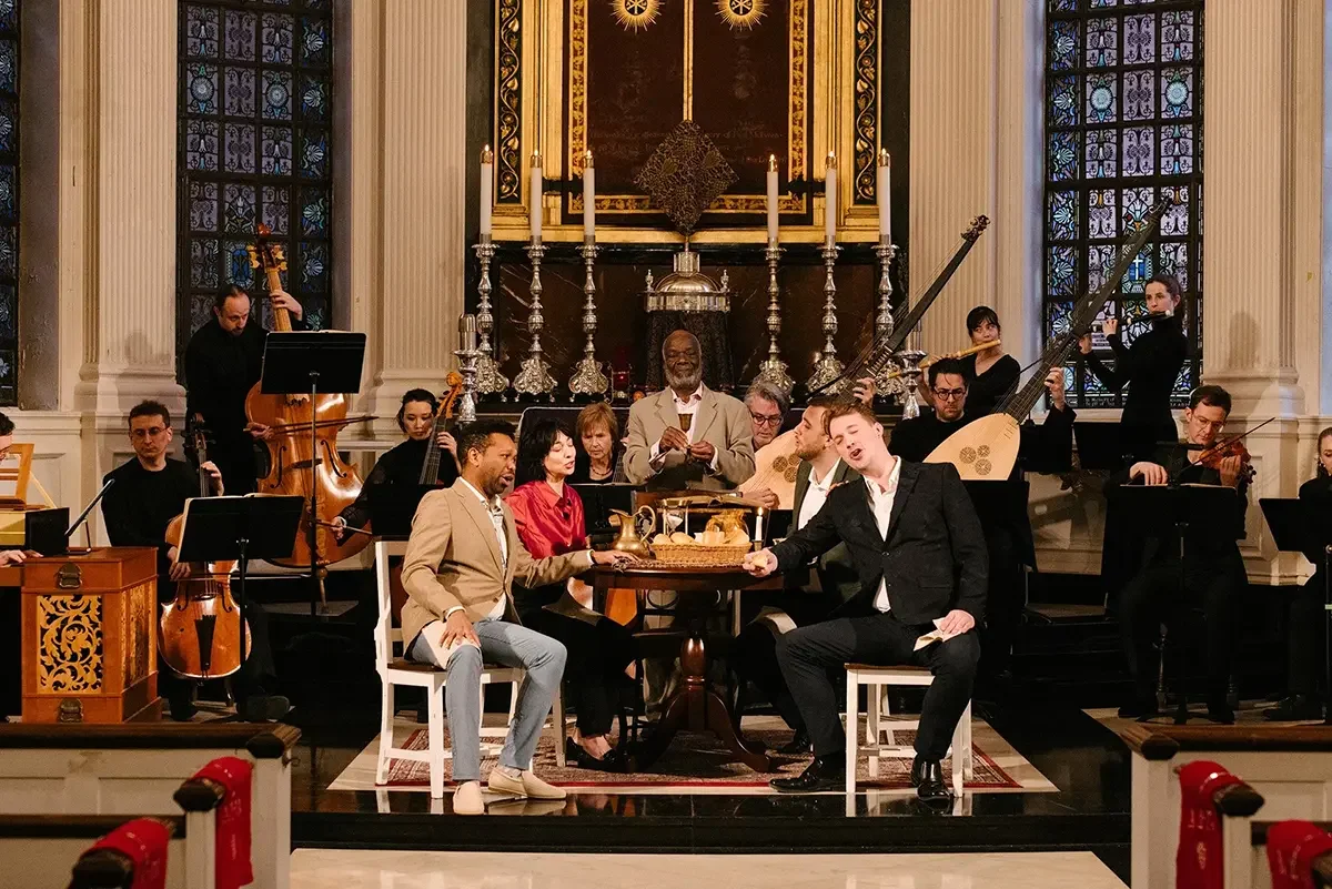 A diverse group of musicians and singers perform in a church sanctuary, surrounded by stained glass windows and ornate candles on a marble altar, with a man in a beige suit and a woman in red seated at a table in the center.