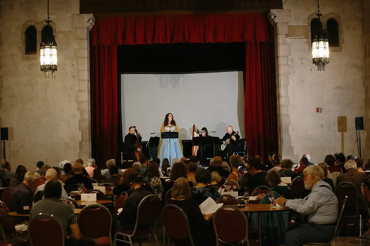 A live musical performance on stage in a large hall with audience members seated at tables. A woman singer is center stage, with a cello player, a harpist, and a guitarist accompanying her. The stage has red curtains and ornate lighting fixtures.