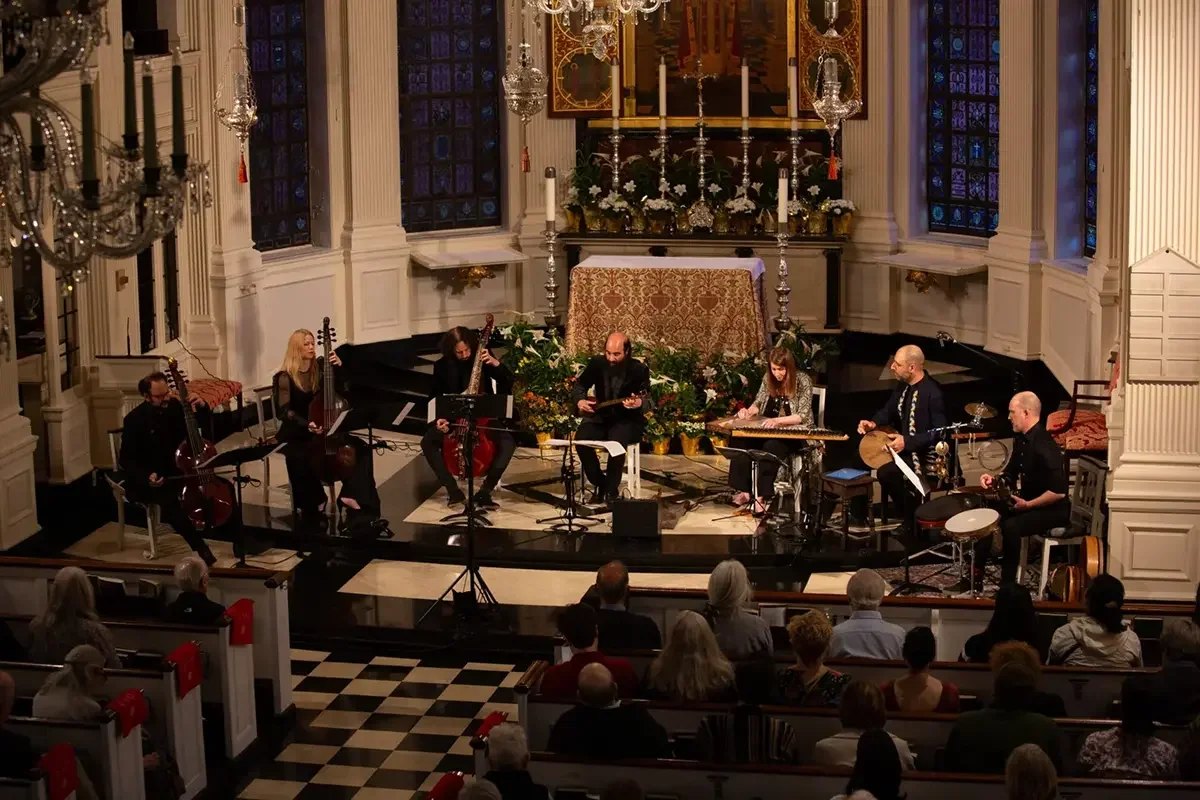 A musical ensemble performing on stage in an ornate church or concert hall with white paneling, stained glass windows, and decorative chandeliers. Audience members are seated in the foreground.