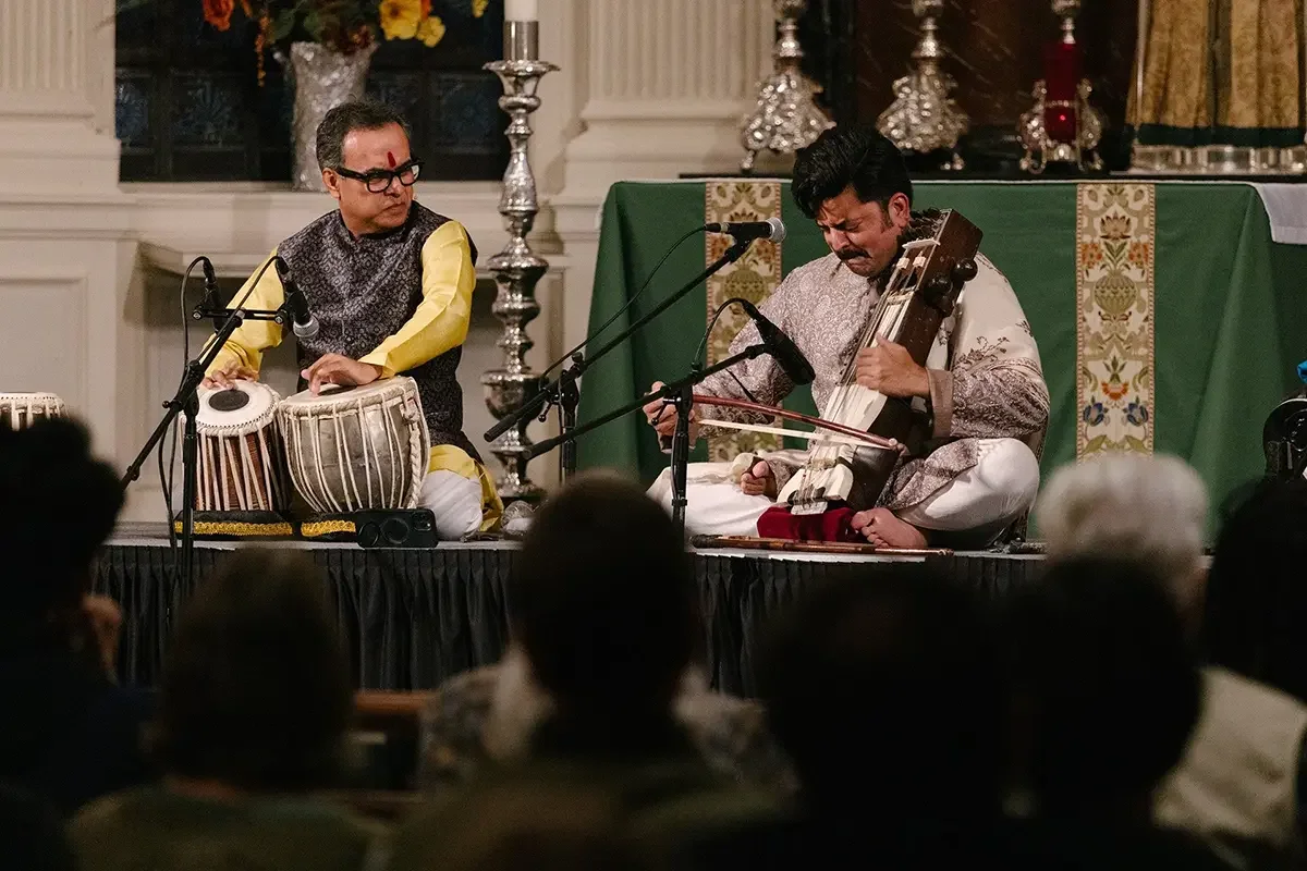 Two musicians performing on stage; one playing tabla drums, the other playing a string instrument, in a decorated indoor setting.