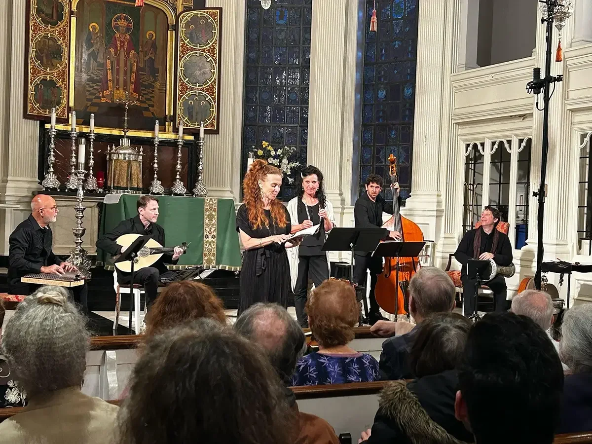 A musical performance inside a church with a group of musicians, including a woman singing and playing a keyboard, and a man playing a double bass. The church features high stained glass windows, a large religious painting, and ornate decor.