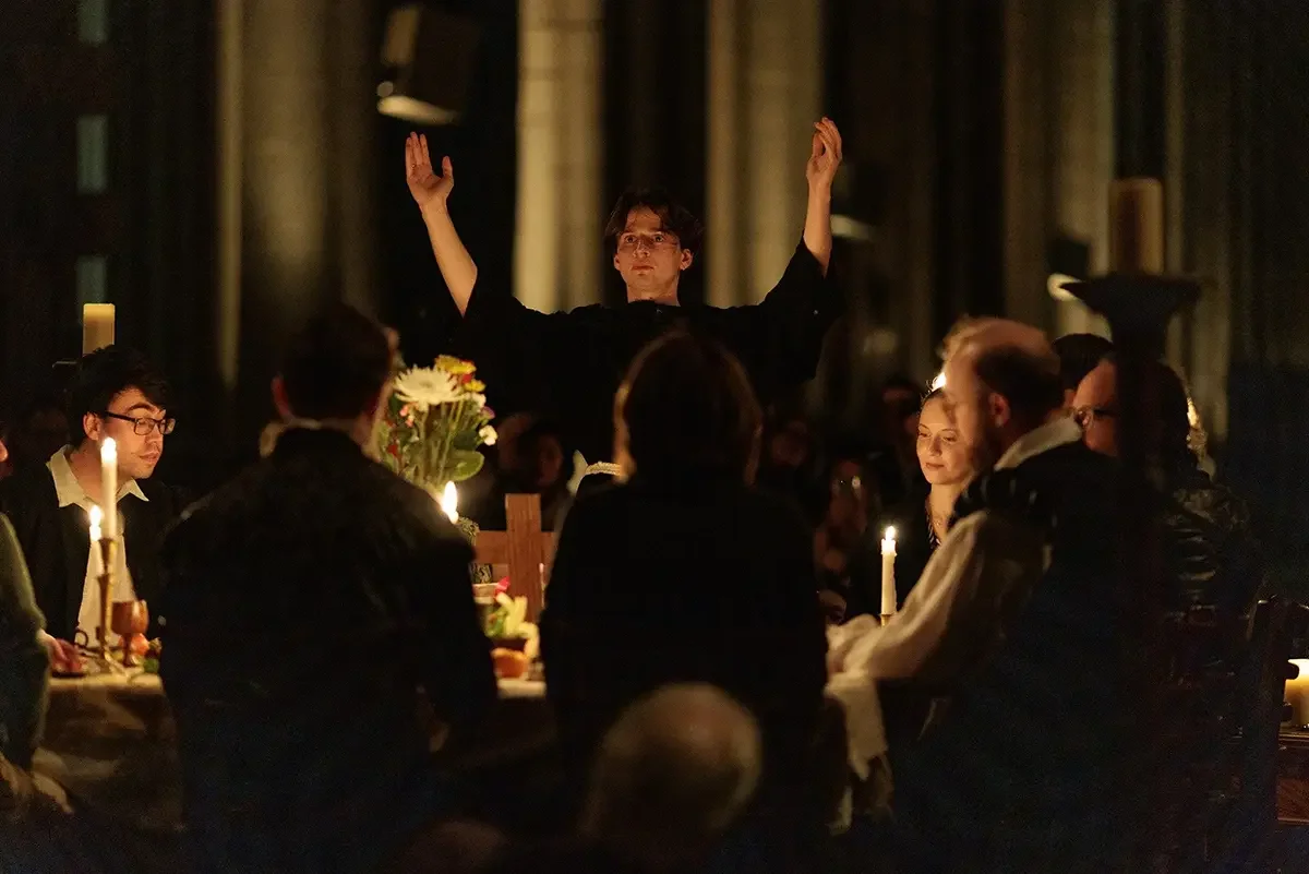 A young man in black robes leads a religious ceremony with raised arms, surrounded by people sitting at a long table with lit candles, flowers, and religious symbols in a dimly lit room.
