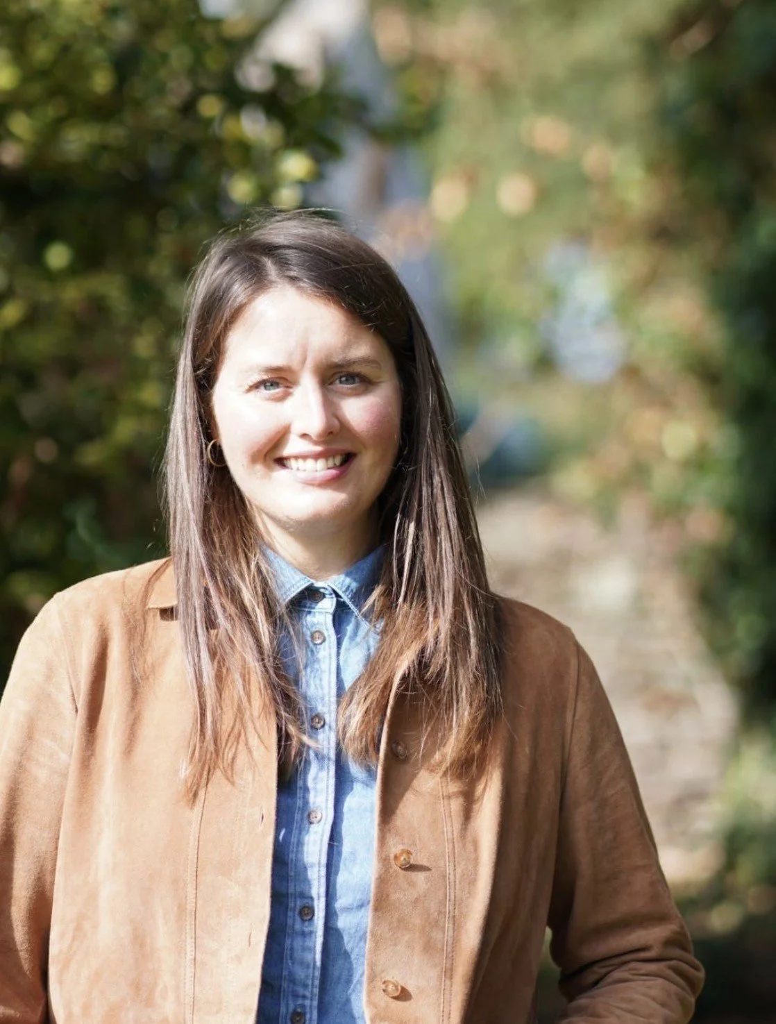 A woman with long brown hair and blue eyes standing outdoors on a sunny day, smiling, with greenery and a dirt path in the background.