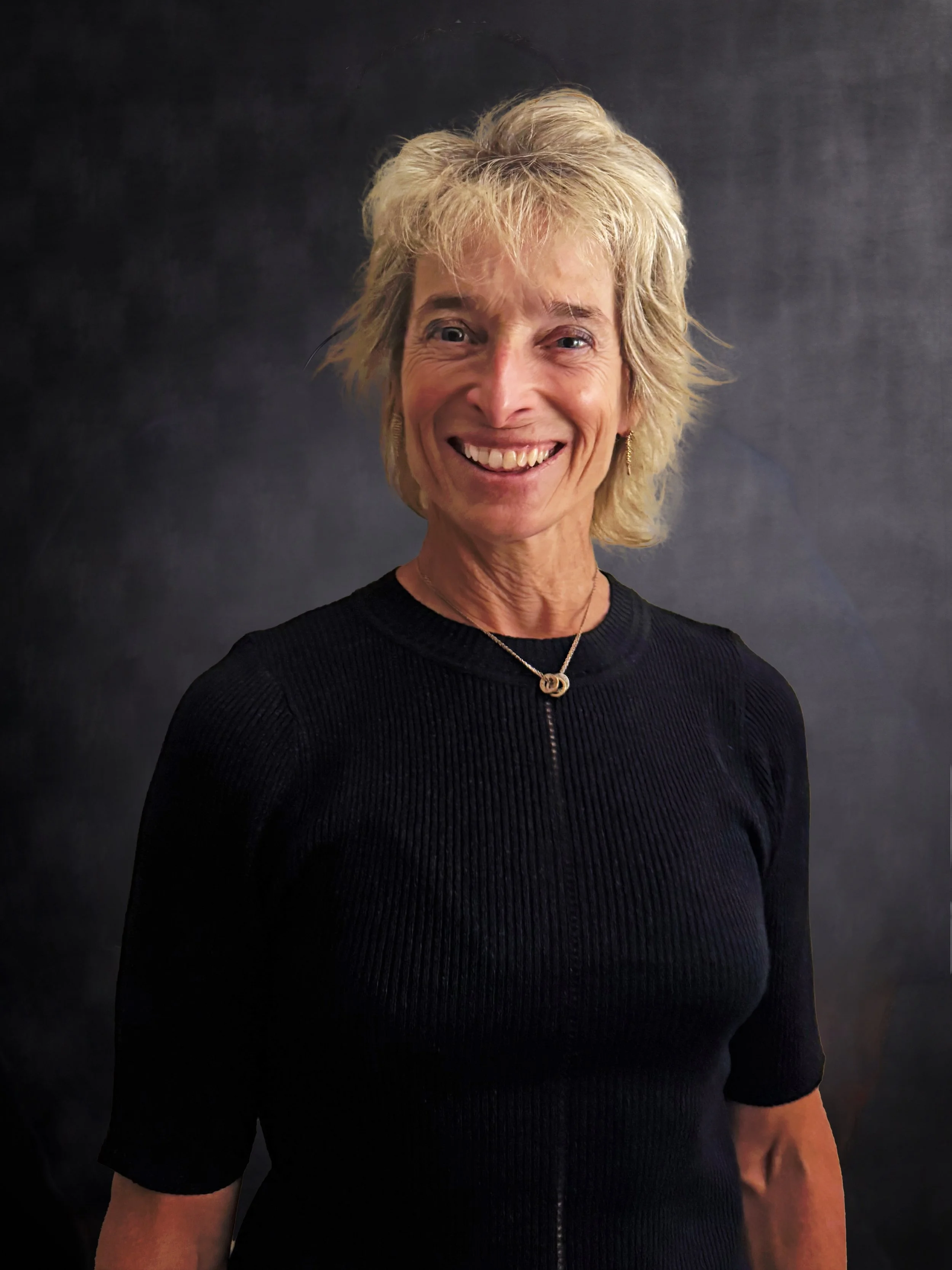 Mulher sorridente com cabelo loiro, cortado na altura dos ombros, usando uma blusa preta de tecido texturizado e um colar com pingente em formato de laço. Fundo escuro de estúdio..