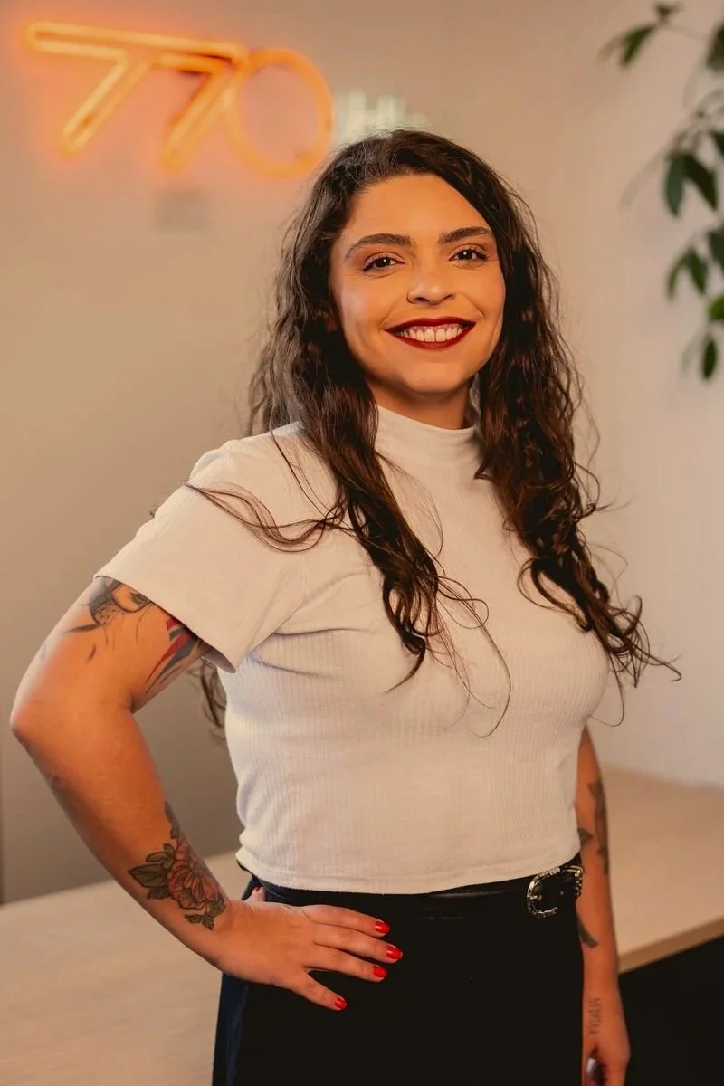 Beatriz, mulher sorridente com cabelos morenos escuros, de largura média, usando uma blusa branca.No fundo, "770" escrito em NEON na parede.