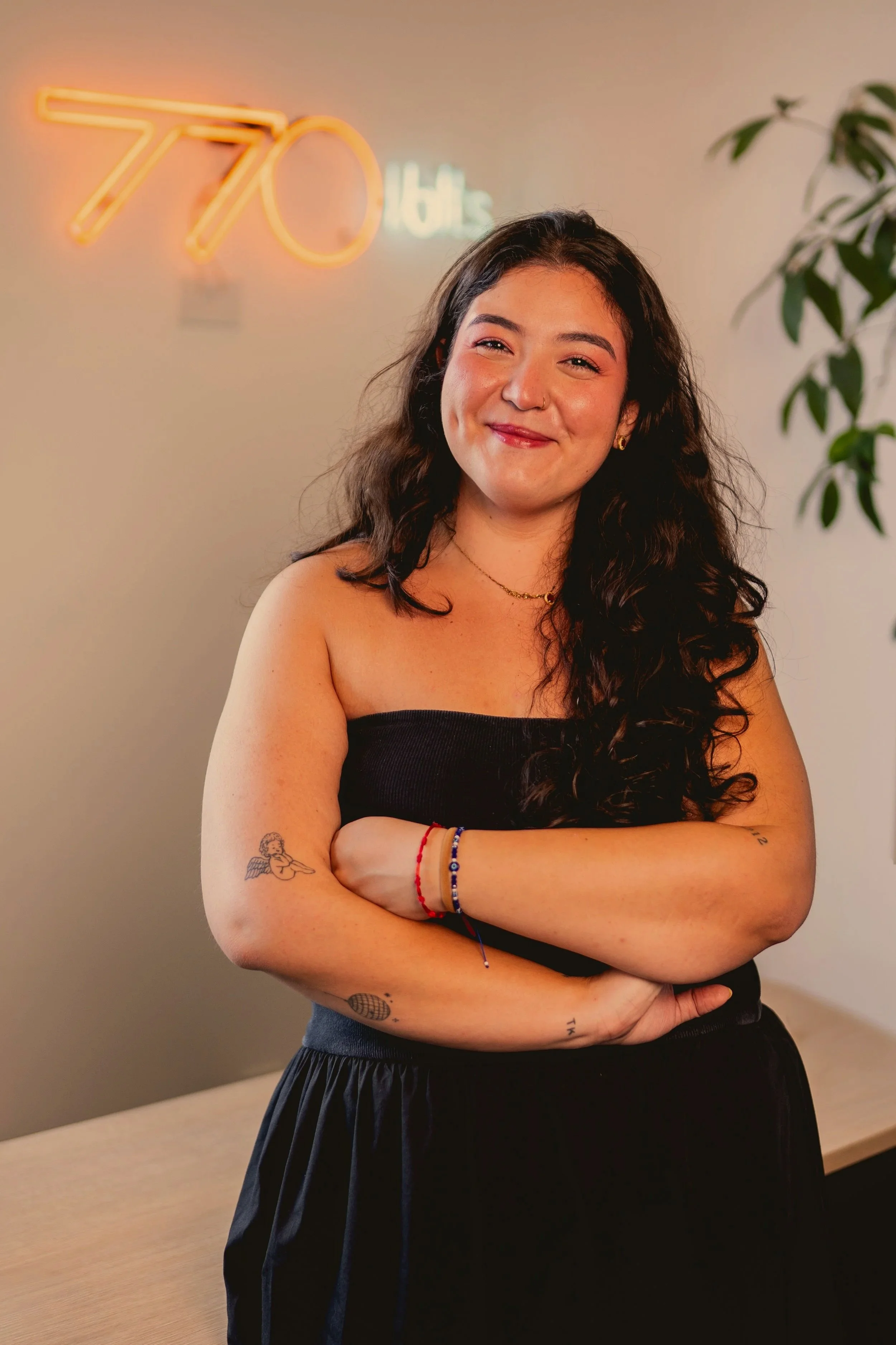 Giovana, mulher sorridente com cabelos longos de cor escura, usando vestido preto, posando com os braços cruzados. No fundo, "770" escrito em NEON na parede.