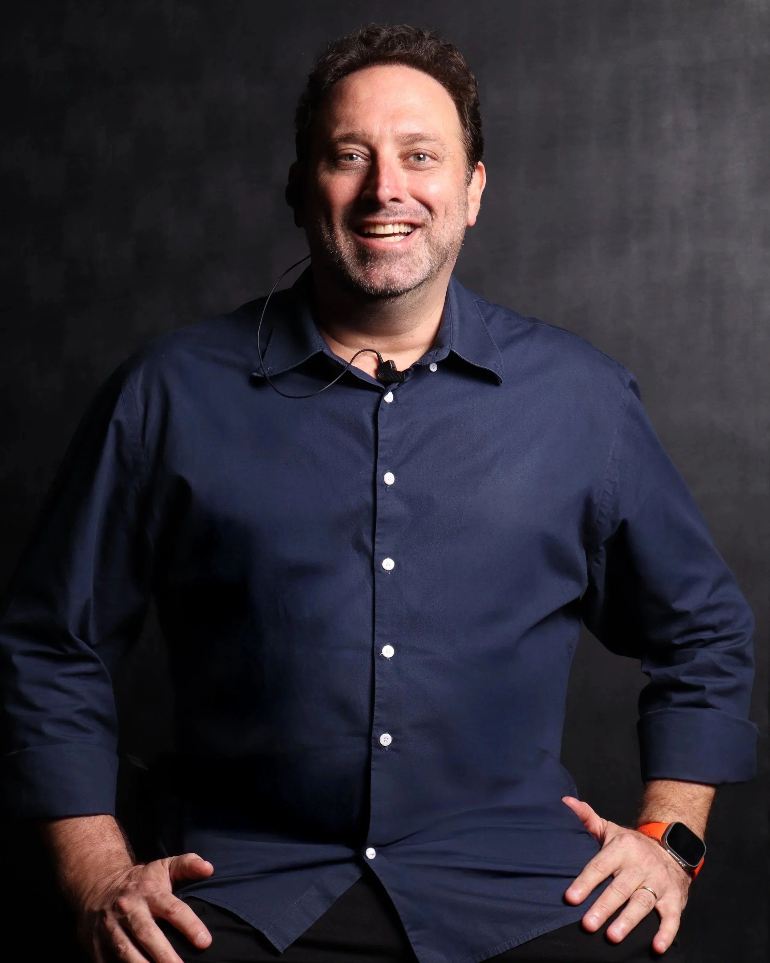 Andreas, homem sorridente usando uma camisa azul escura com um fundo escuro de estúdio.