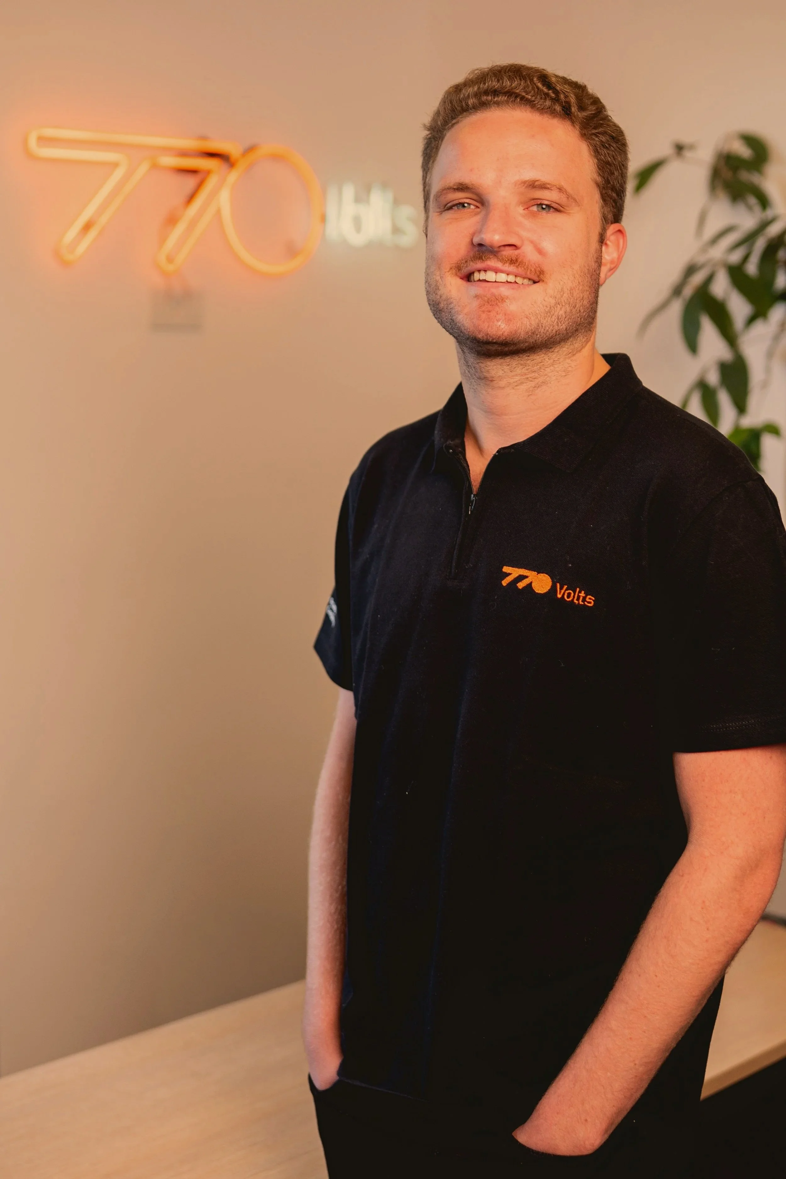 Pablo, homem sorridente com cabelo cacheado de cor loira/morena, barba leve, polo preta com "770 Volts" escrito, contra fundo claro com "770" em neon.