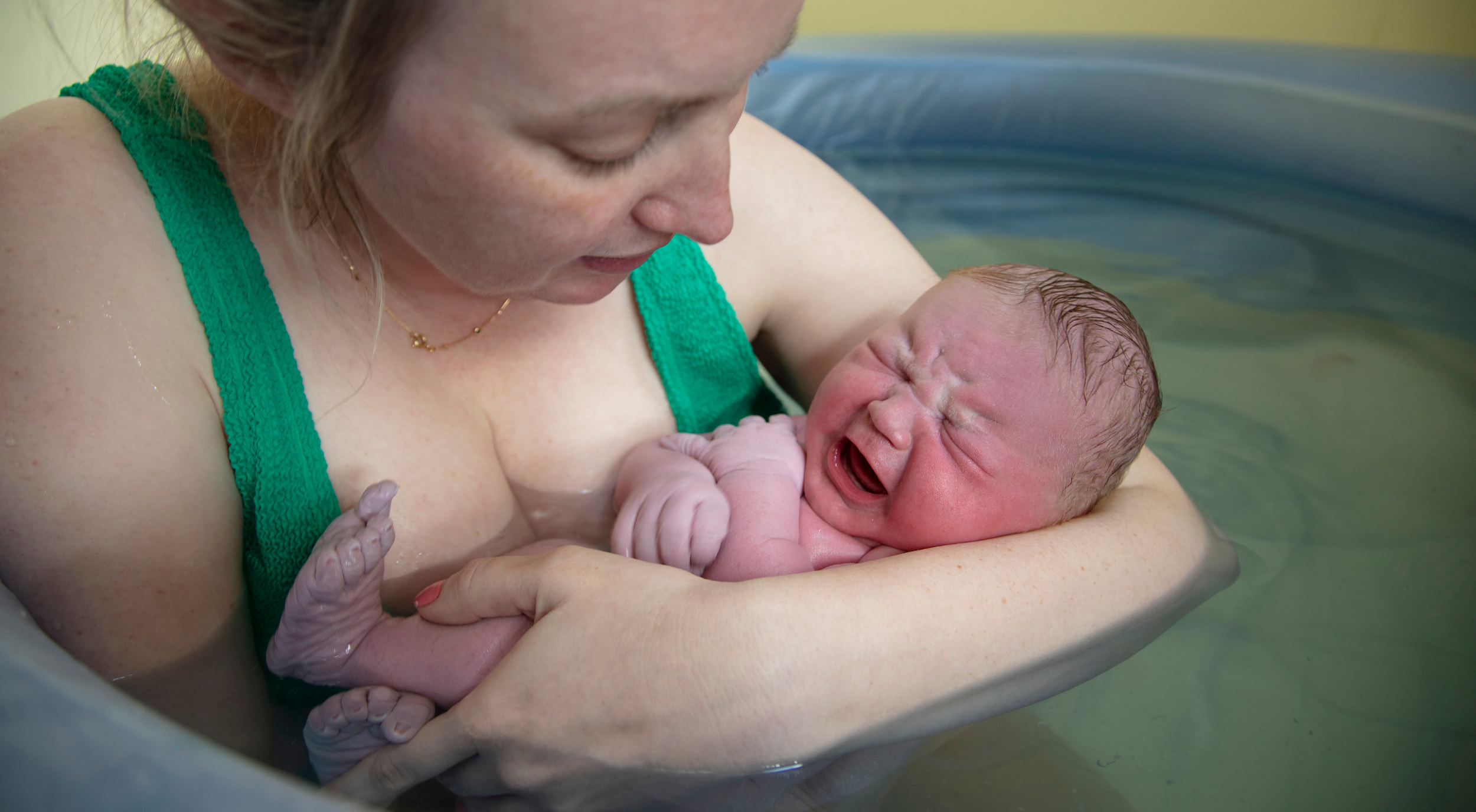 A woman holds a crying newborn baby in a blue infant bathtub, with water surrounding them.