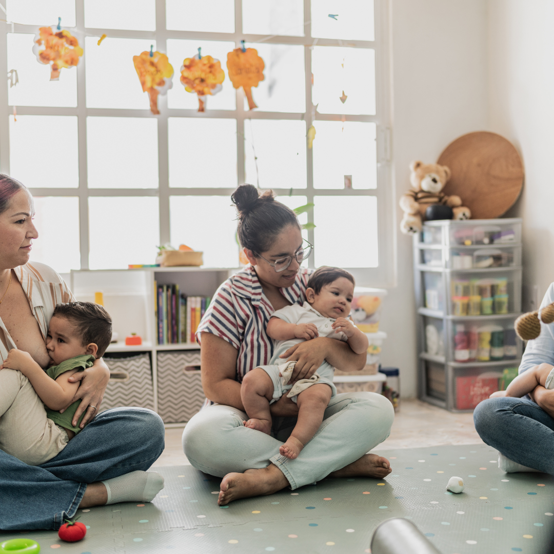 Group of women with young children sitting on a colorful mat in a well-lit playroom. One woman holds a baby, another woman cradles a toddler, and the third woman is partially visible, holding her child. Play and educational supplies are visible in the background.