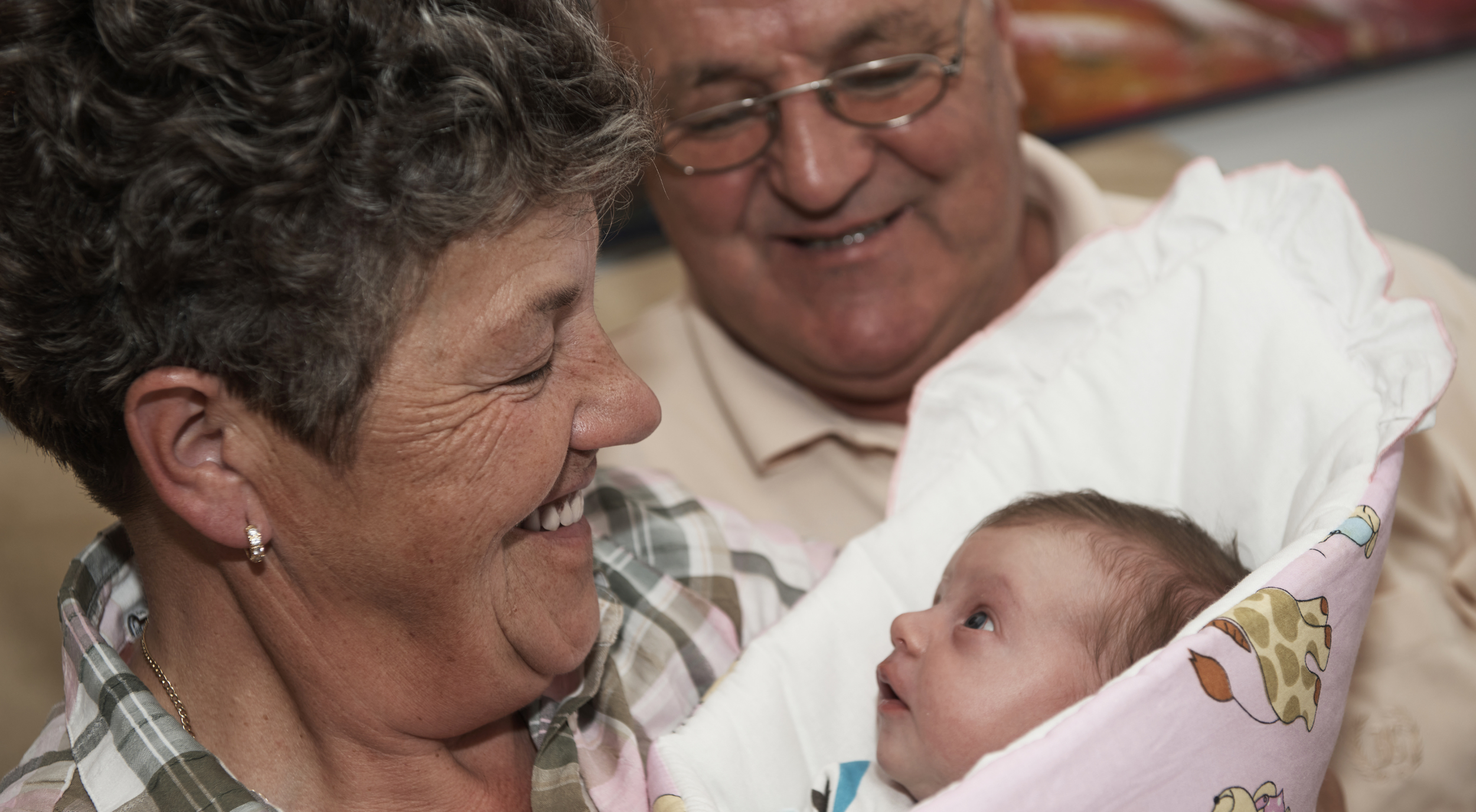 An elderly woman holding a baby in a cradle, smiling at the baby while an elderly man looks at the baby and smiles.