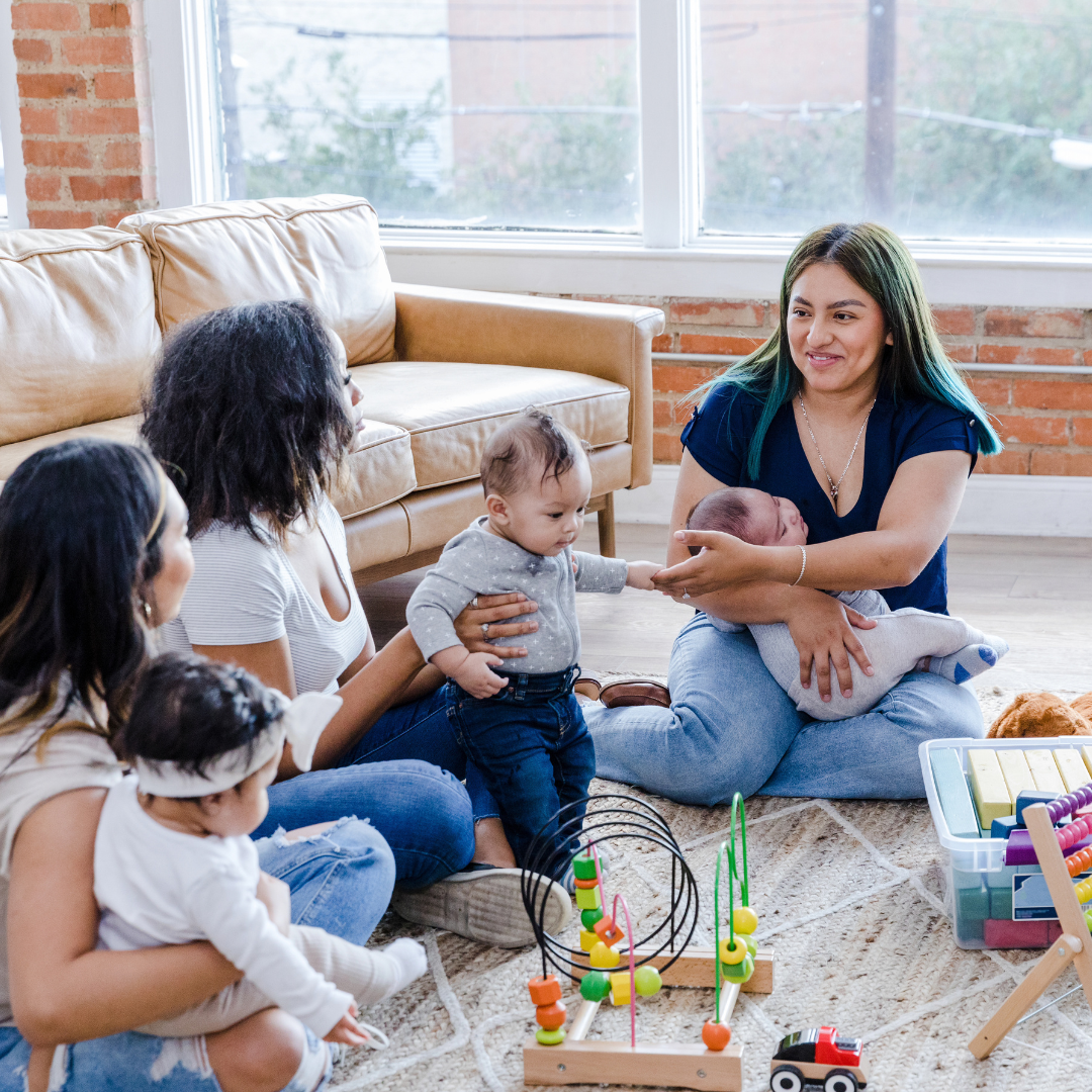 Women and children in a living room with a couch and large window, one woman holding a newborn, another woman with a toddler reaching out, with toys on the carpet.