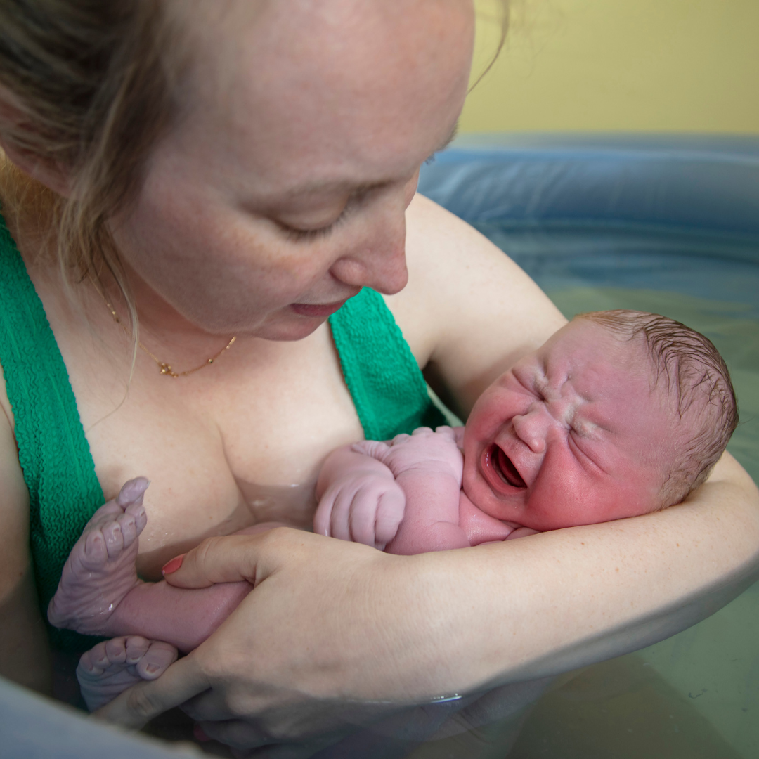 A woman holding a newborn baby during a water birth in a birthing tub, both looking at each other.