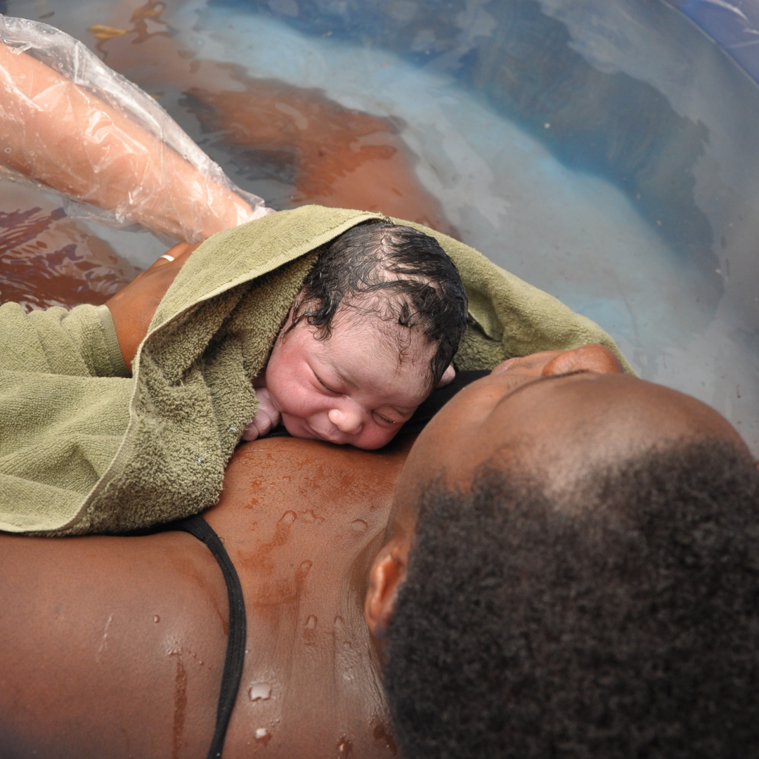 A newborn baby is being delivered via water birth, lying on a person's chest in a birthing pool. The baby is wrapped in a green towel and is resting its head on the adult's chest, with closed eyes and wet hair. The parent is partially submerged in water, with water droplets on their skin, and it is a moment of close bonding.
