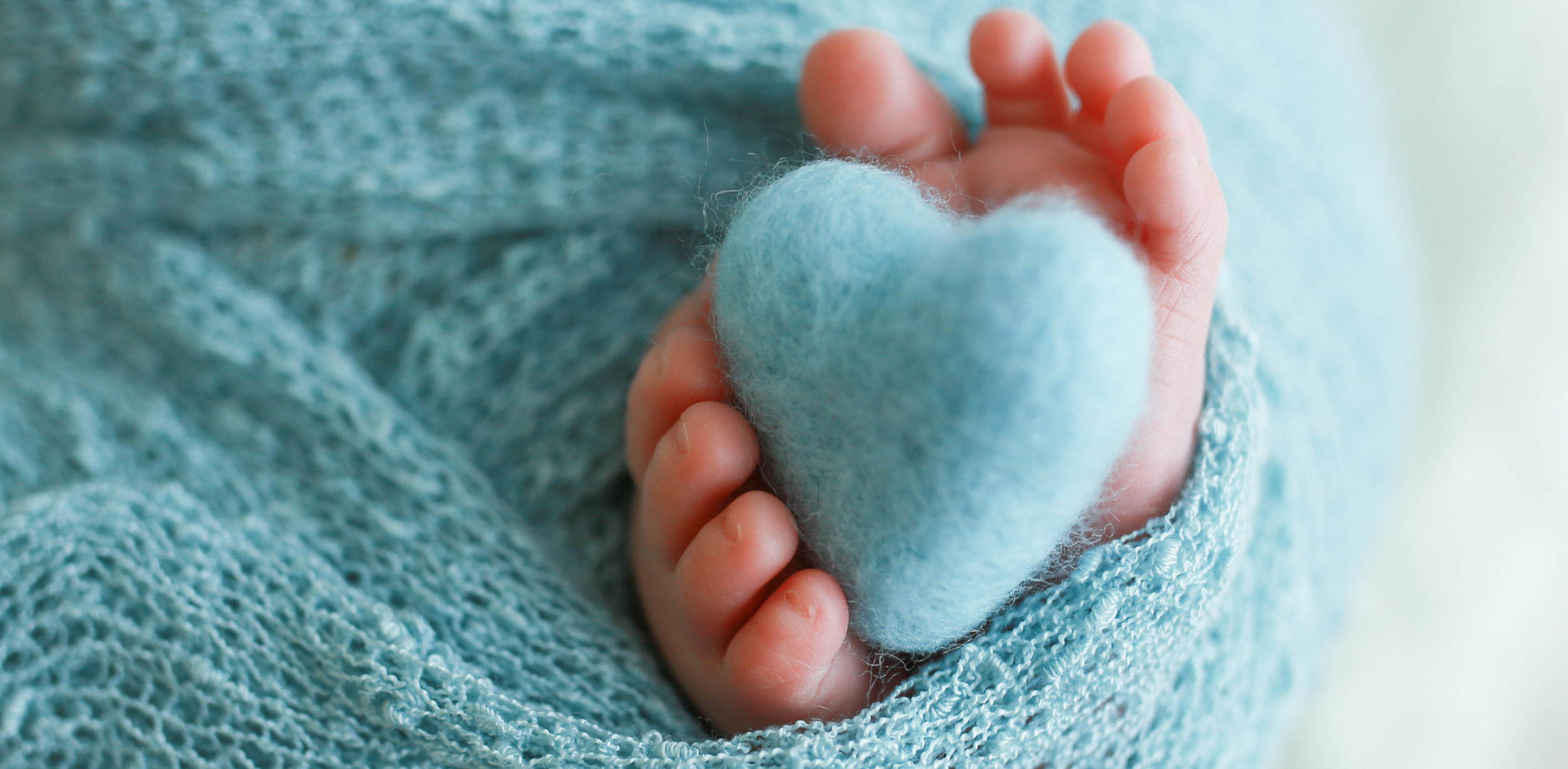 A close-up of a tiny baby's feet holding a soft, light blue heart-shaped object, with the baby's hand and object wrapped in a textured blue fabric.