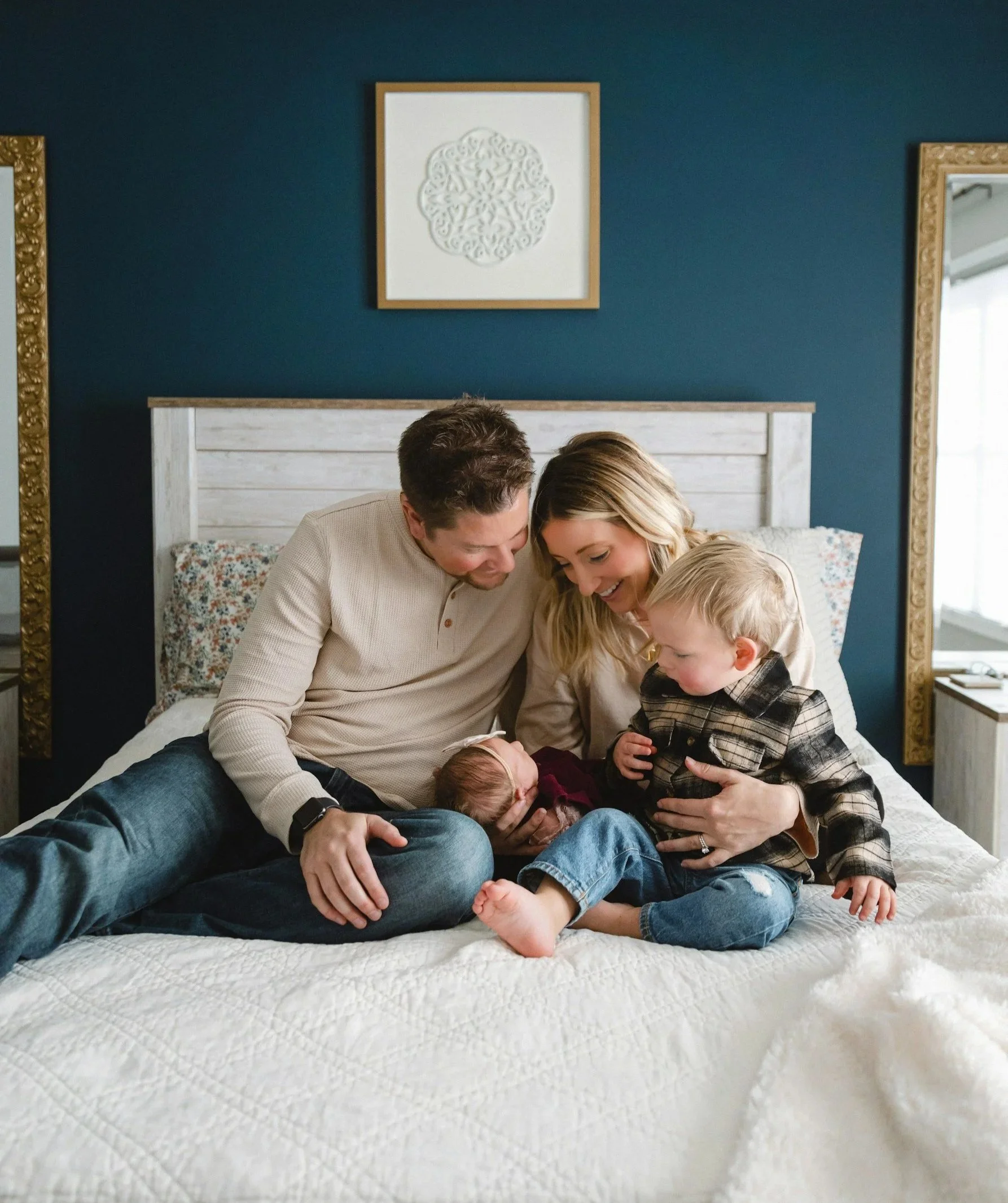 A family of four sitting on a bed, sharing a joyful moment with their newborn, who is being held by the mother. The father and older child are looking at the baby, smiling.