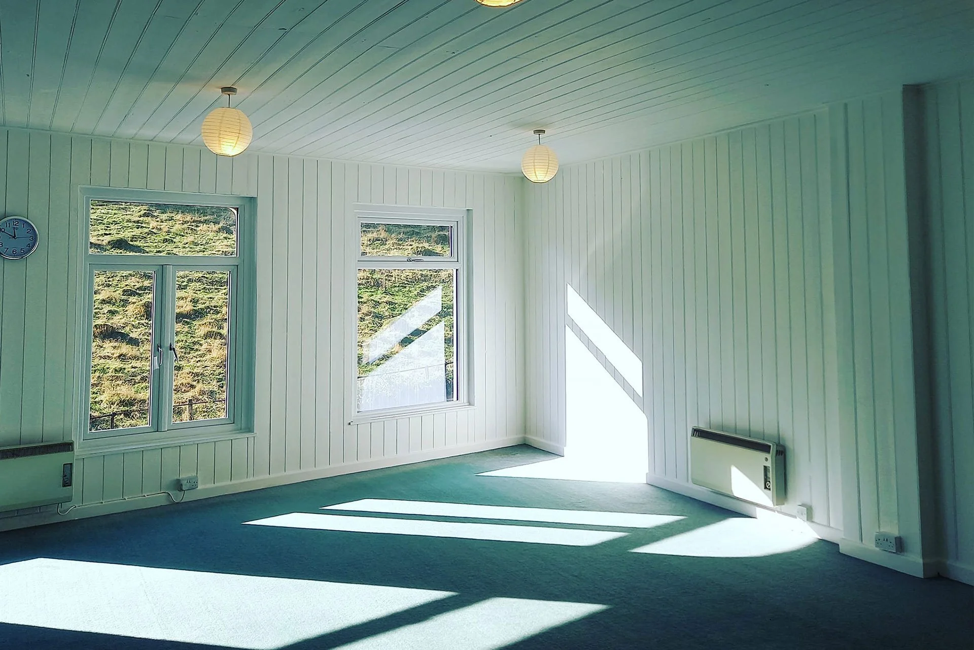 Empty room with white wooden panel walls, large windows letting in sunlight, and ceiling-mounted paper lanterns. Green carpet on the floor, with shadows cast by the sunlight.
