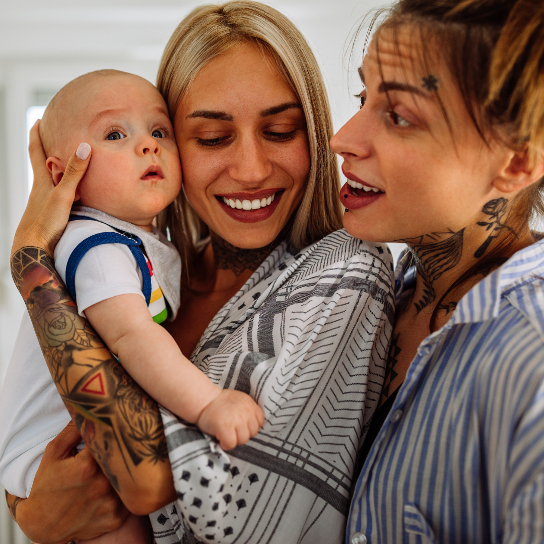 Three people, two women with tattoos, and a baby, smiling and holding each other in a close embrace indoors.