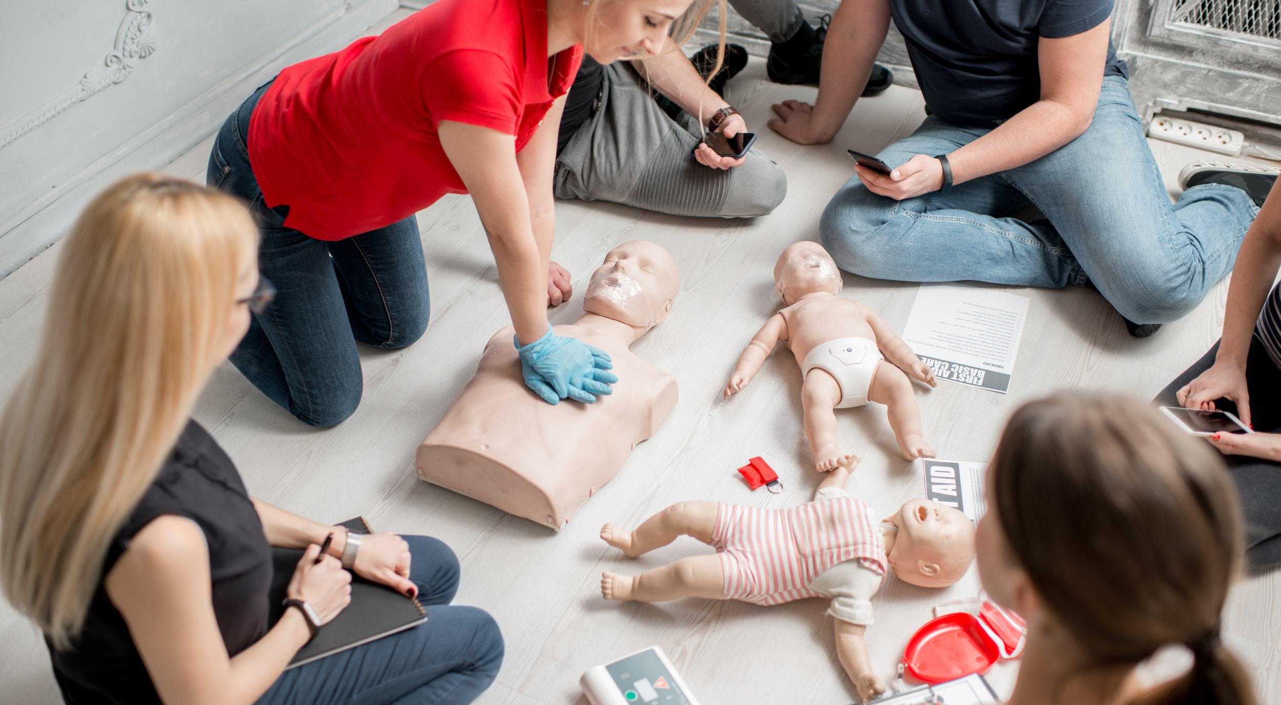 Group of people attending a CPR training session, practicing on baby and adult mannequins, with medical supplies and instructions on the floor.