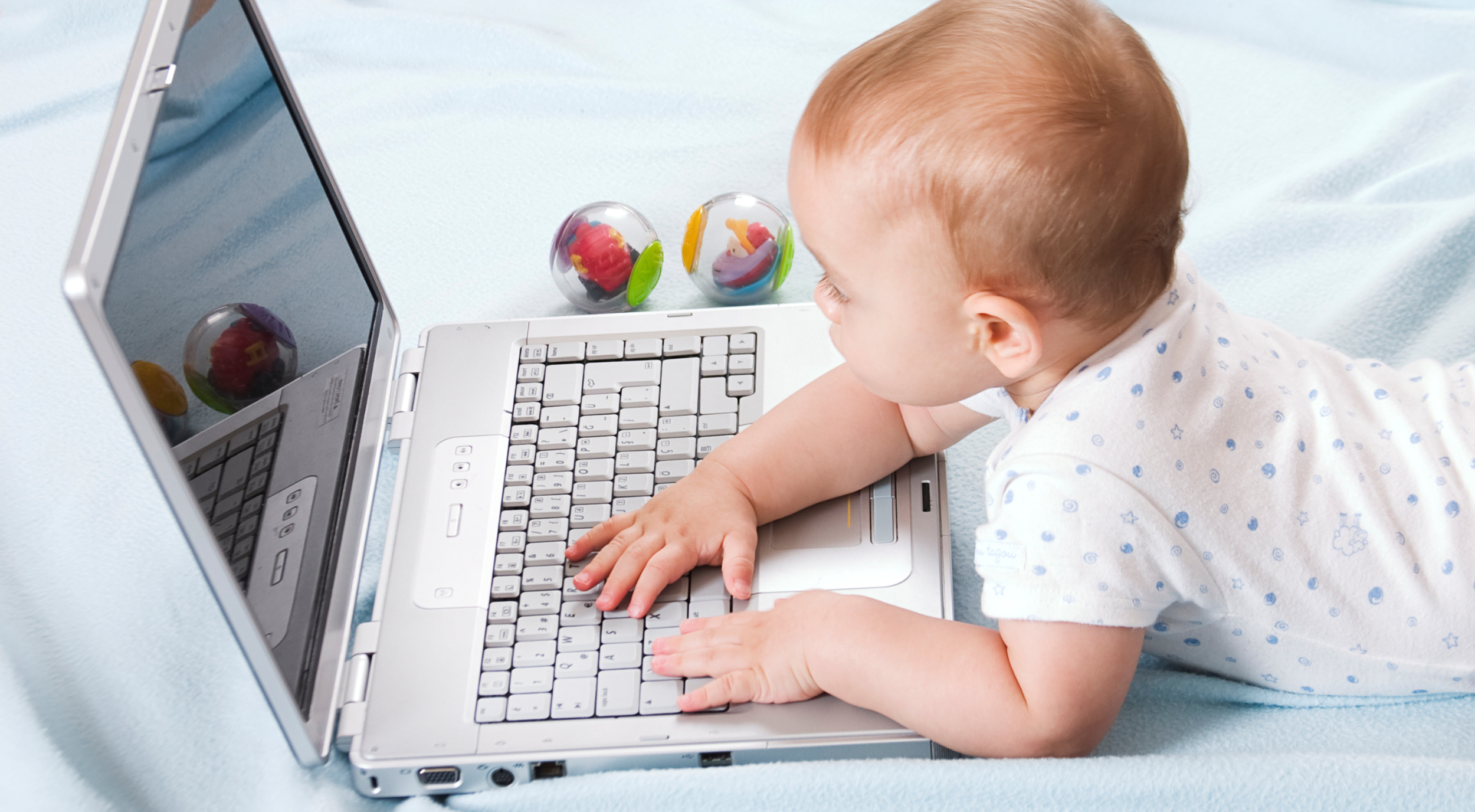 A baby lying on a light blue blanket, looking at a laptop computer with colorful plastic balls nearby.